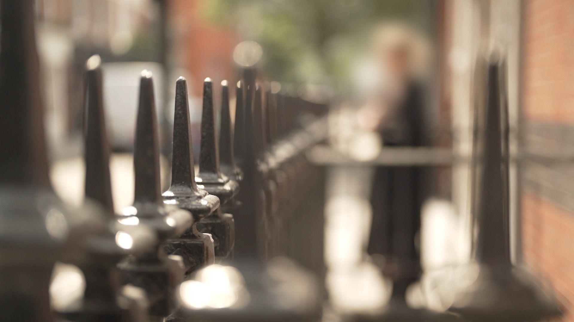 Emma stands outdoors beside a brick building, deliberately out of focus to conceal her identity. A row of sharply focused black metal railings runs through the foreground.