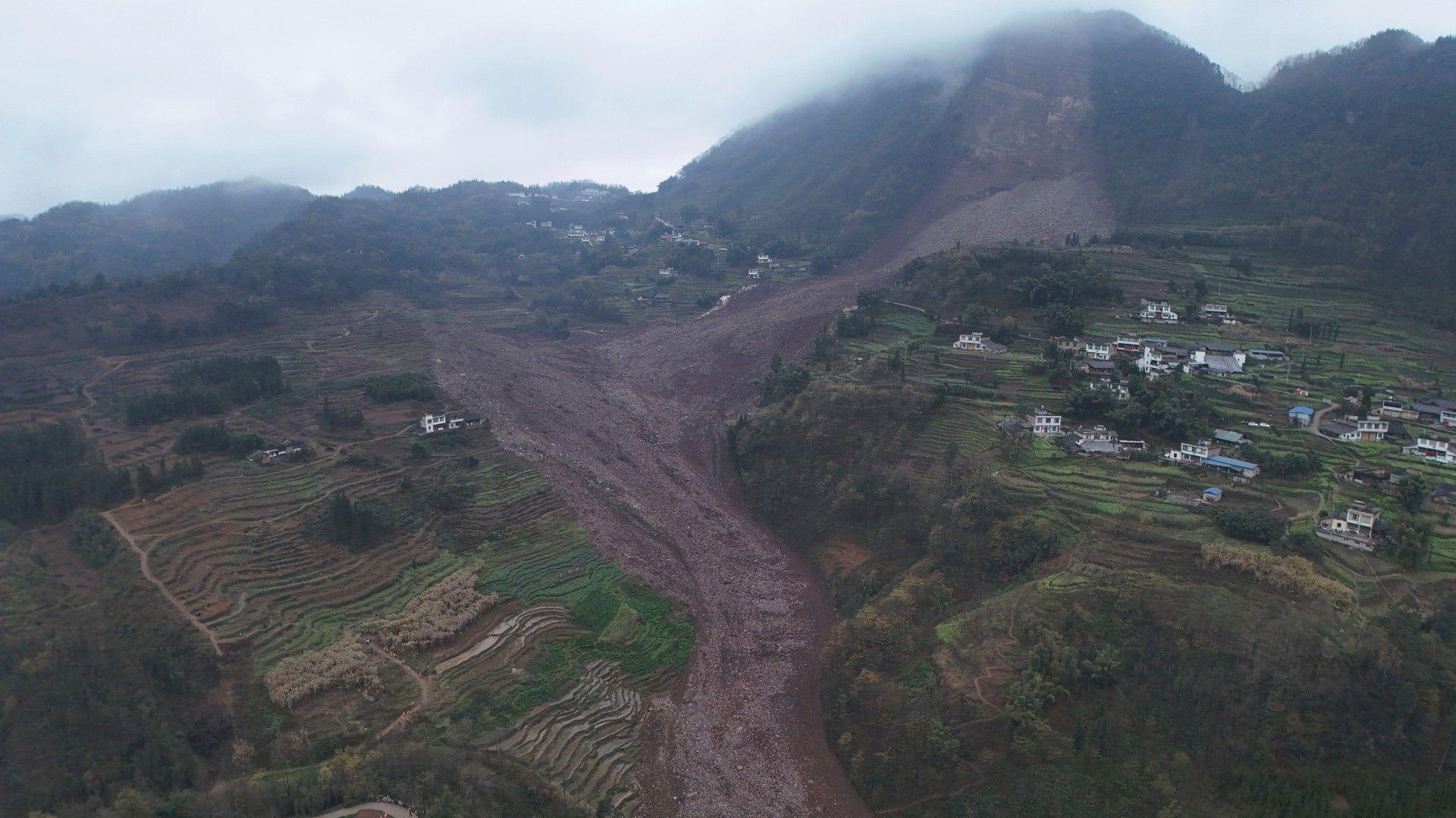 An aerial shot shows the damage after a landslide in south-western China. A large brown slash of mud and rock cuts through the otherwise green landscape.