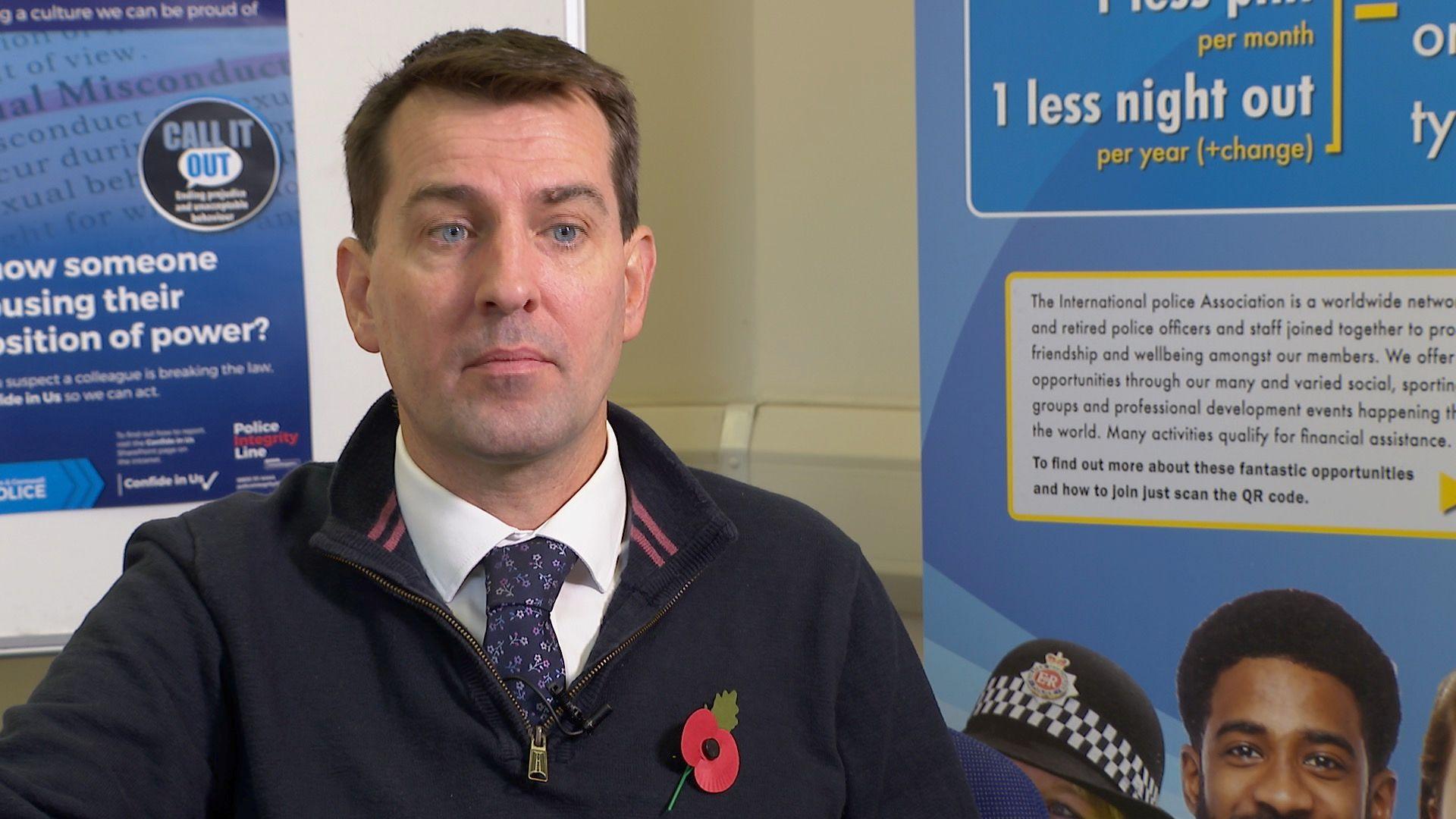 Man with short brown hair and blue eyes looks at the camera while wearing a half zip top with shirt and flower pattern tie visible under and a paper poppy on his left chest. In the background is a white wall and parts of two posters, one showing the top of a police woman's hat.