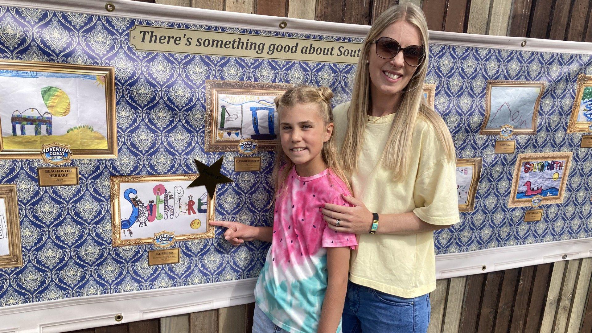 Ellie and her mum stand and point at Ellie's artwork printed on the wall beside her. They both have blonde hair and are smiling. Her mum wears black sunglasses and Ellie is wearing a multicoloured tie-dye t-shirt.