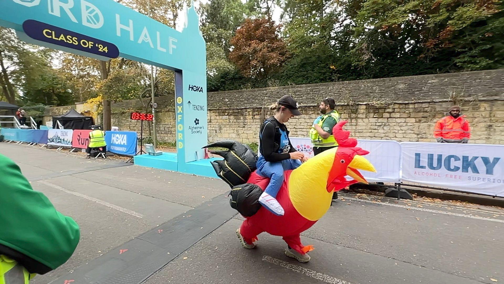 A woman dressed as an enormous chicken - she has just crossed the large blue finish line and is checking her watch.