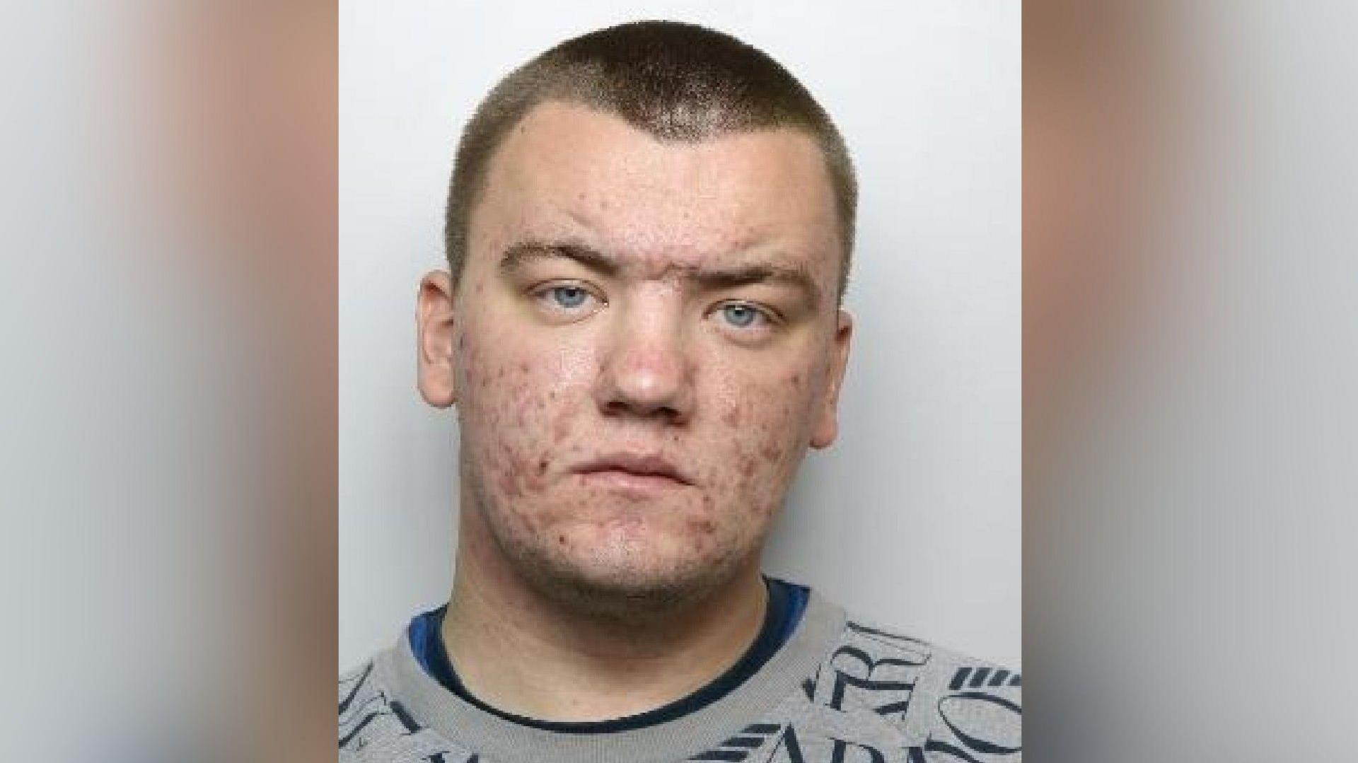 A mugshot of Huggins who has a shaven head and blue eyes. He is wearing a grey top and looks at the camera unsmiling.