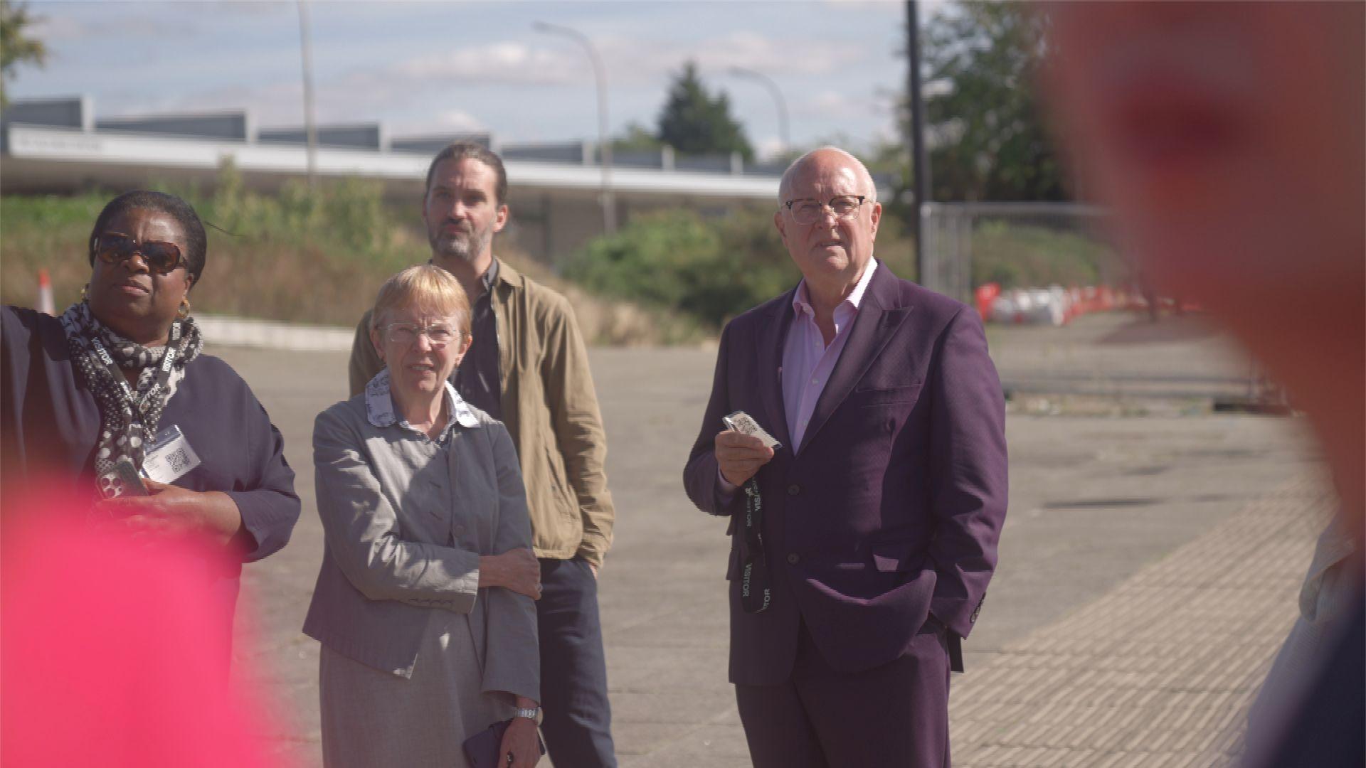 A candid shot from the new towns taskforce's walkabout in Milton Keynes. Four people can be seen in the shot, including Sir Michael Lyons. They are outside, standing on a paved area with a low rise building in the background.