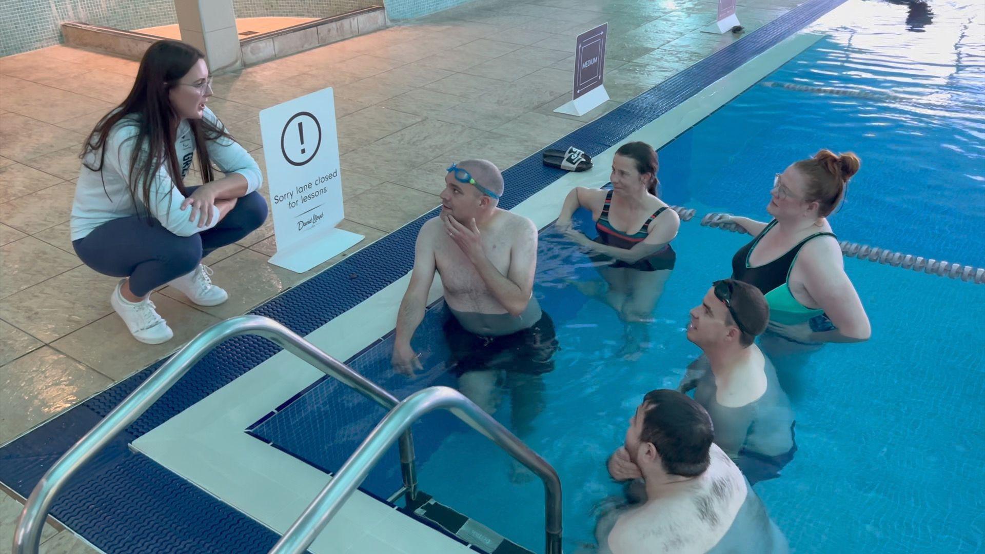 Group of six people at a swimming pool. One person, a woman, is stood crouched on the side of the pool and five people including three men and two women are in the swimming pool.