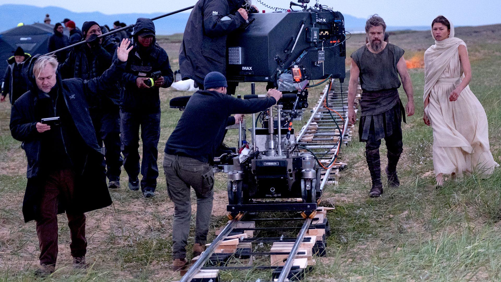 Christopher Nolan directs at an outdoor shoot for Odysseus - he is standing on the left in black clothing, a camera and crew are in the centre of the image and Matt Damon and Zendaya are on the right