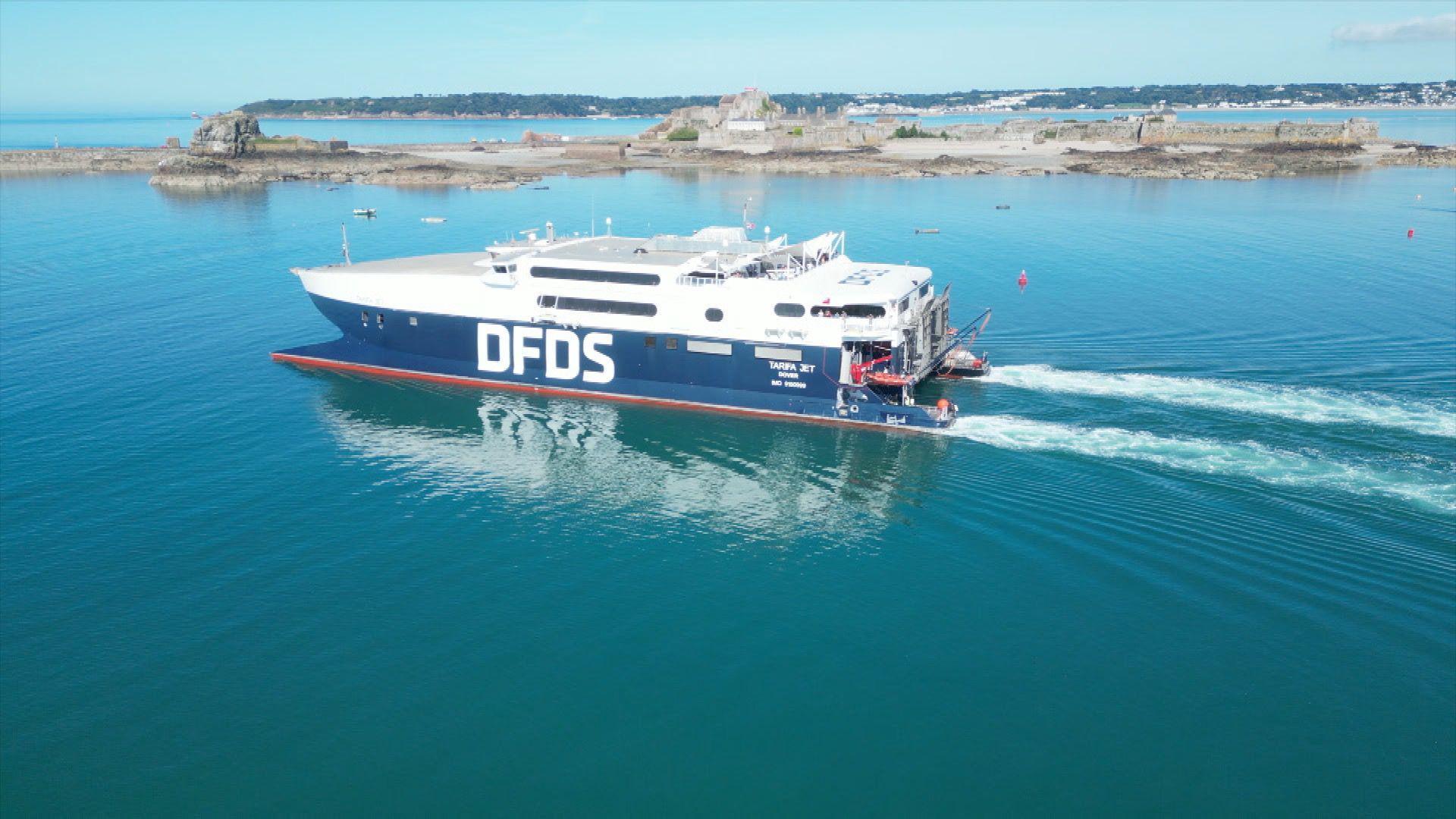 The side of a large white and blue ferry on the water. It is white at the top and blue at the bottom. The water is blue and there is a land in the behind it.
