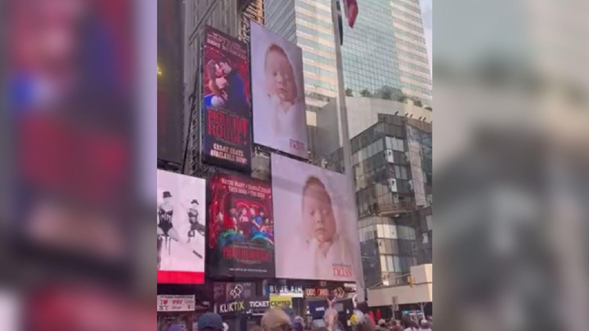 An image of a baby girl with Down's syndrome is screened on advertising display screens in front of a crowd of cheering people. There are skyscrapers in the background and a US flag on a pole.