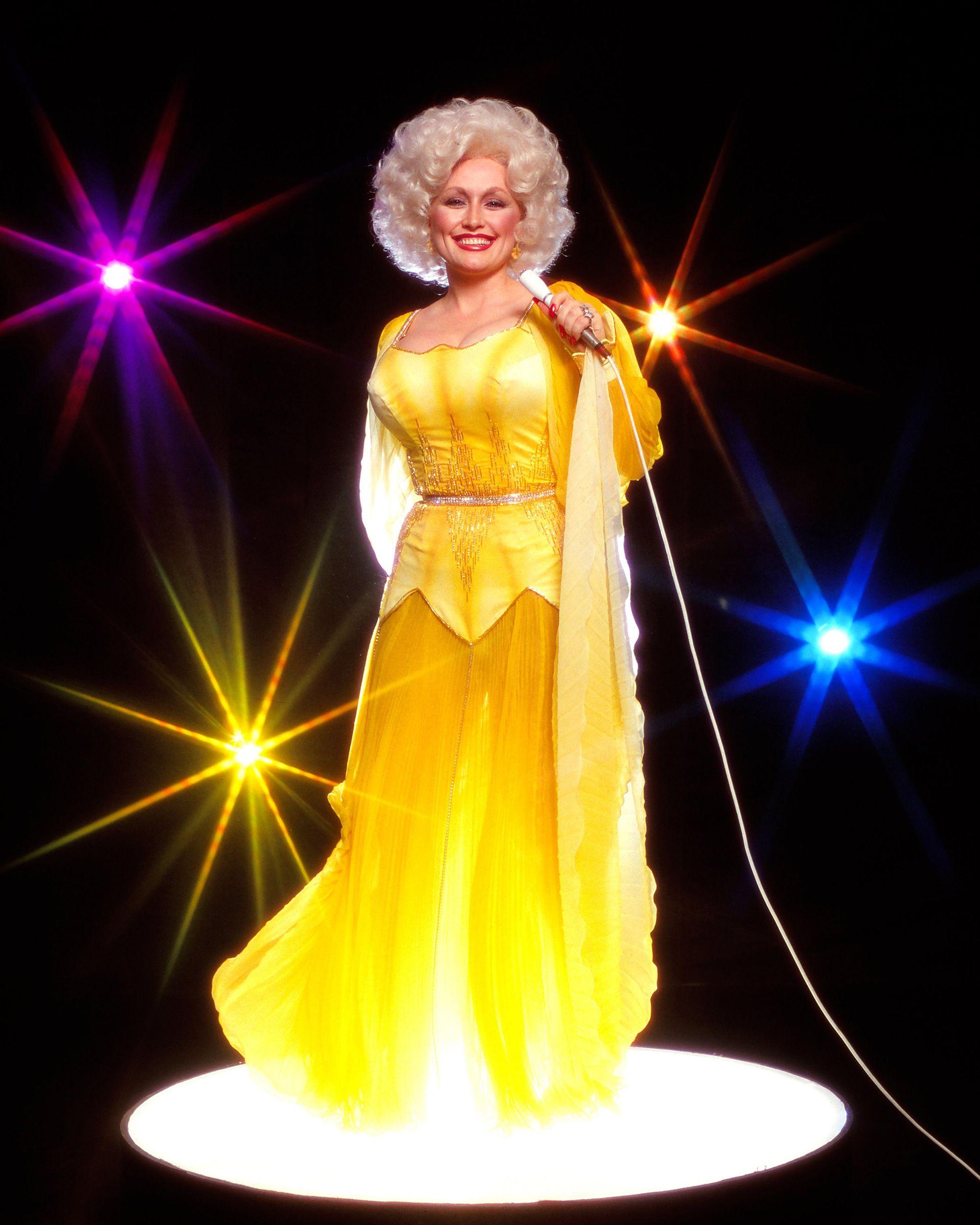 Dolly Parton poses for a portrait session in 1978. She holds a microphone against a black backdrop and wears a bright yellow flowing outfit.