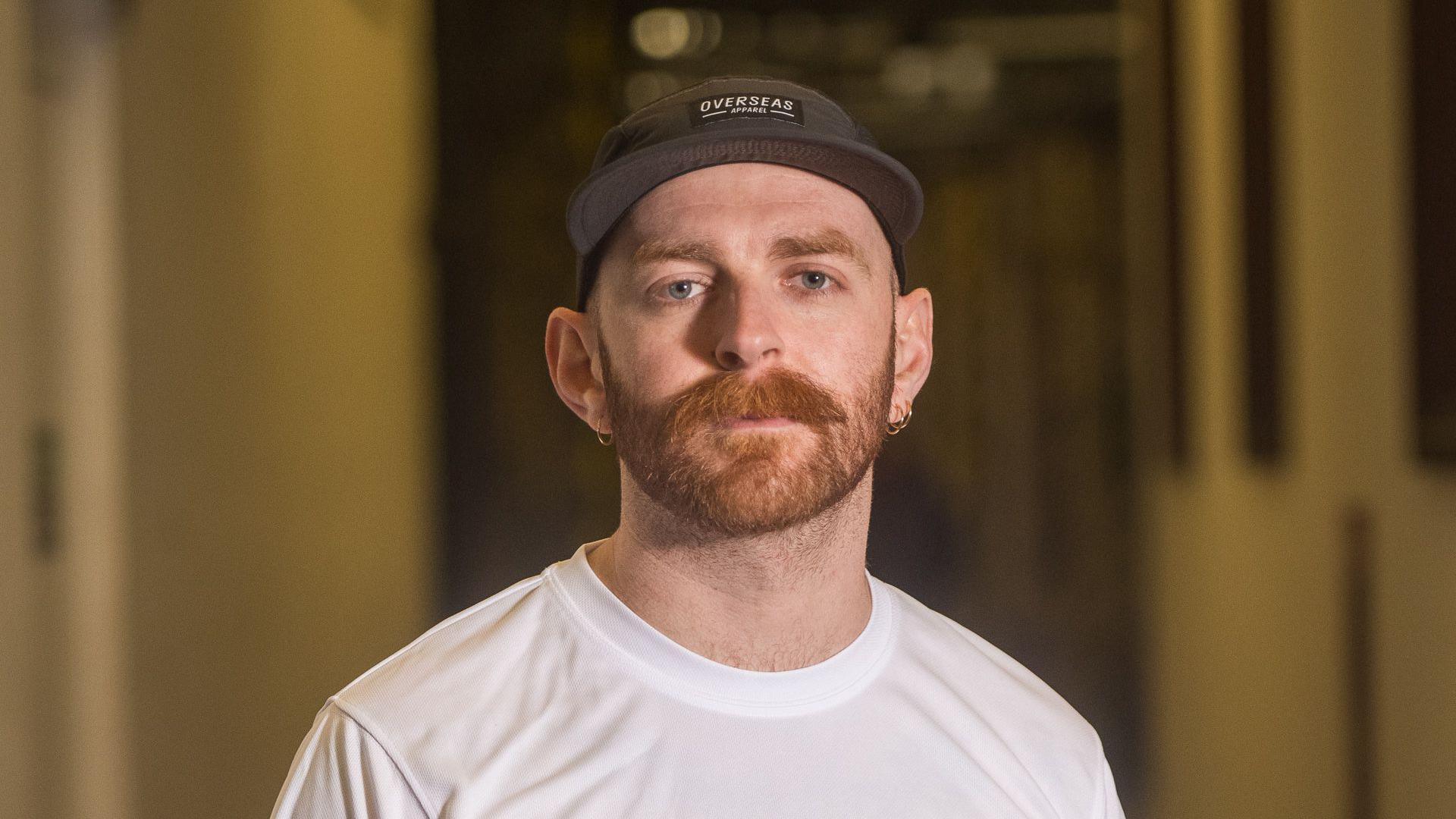 Liam Jones looks at the camera, he has a ginger beard and moustache. He is wearing a black cap and a white t-shirt.