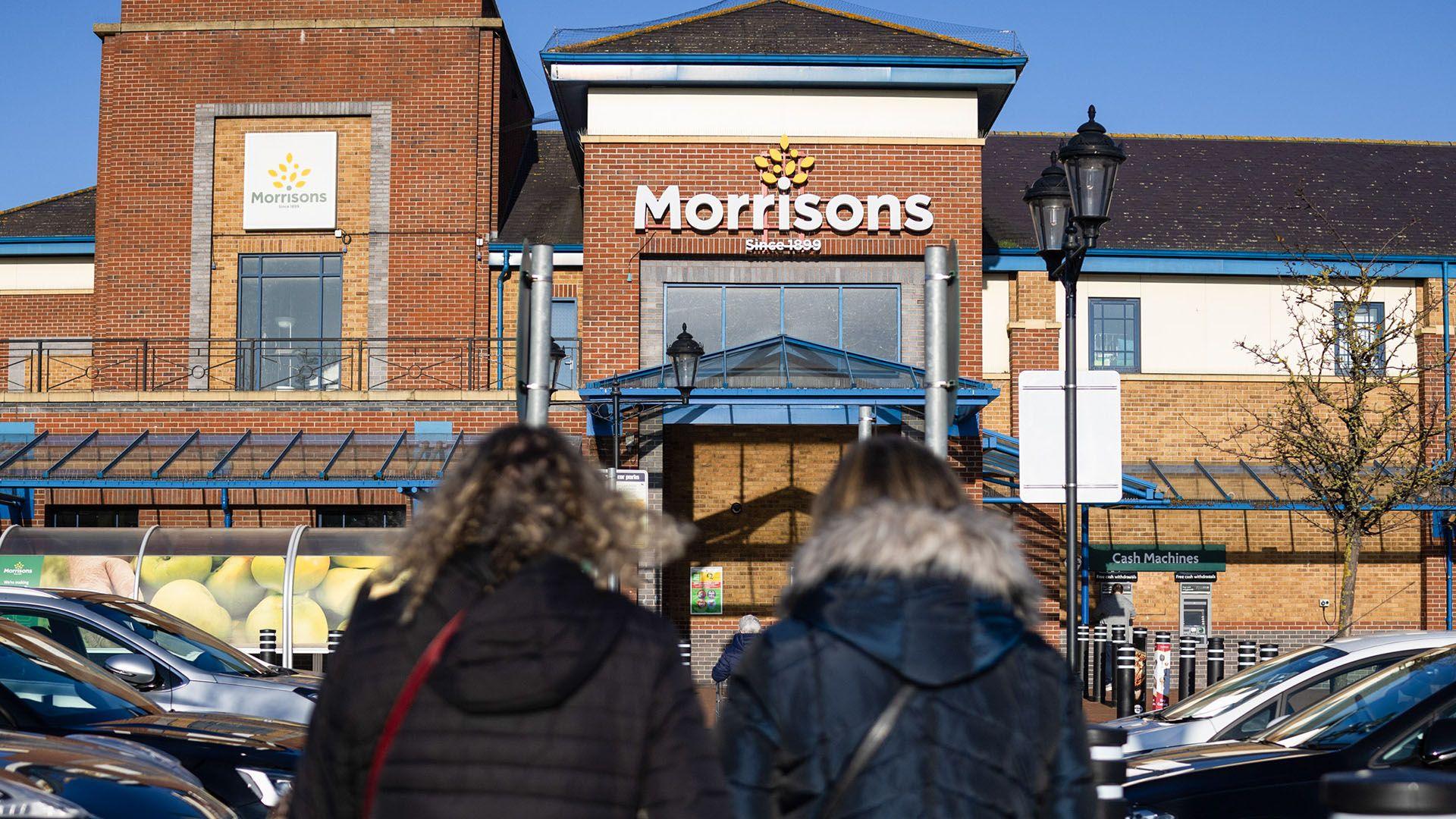 Two women in coats walking into a large Morrisons store with the logo above the entrance