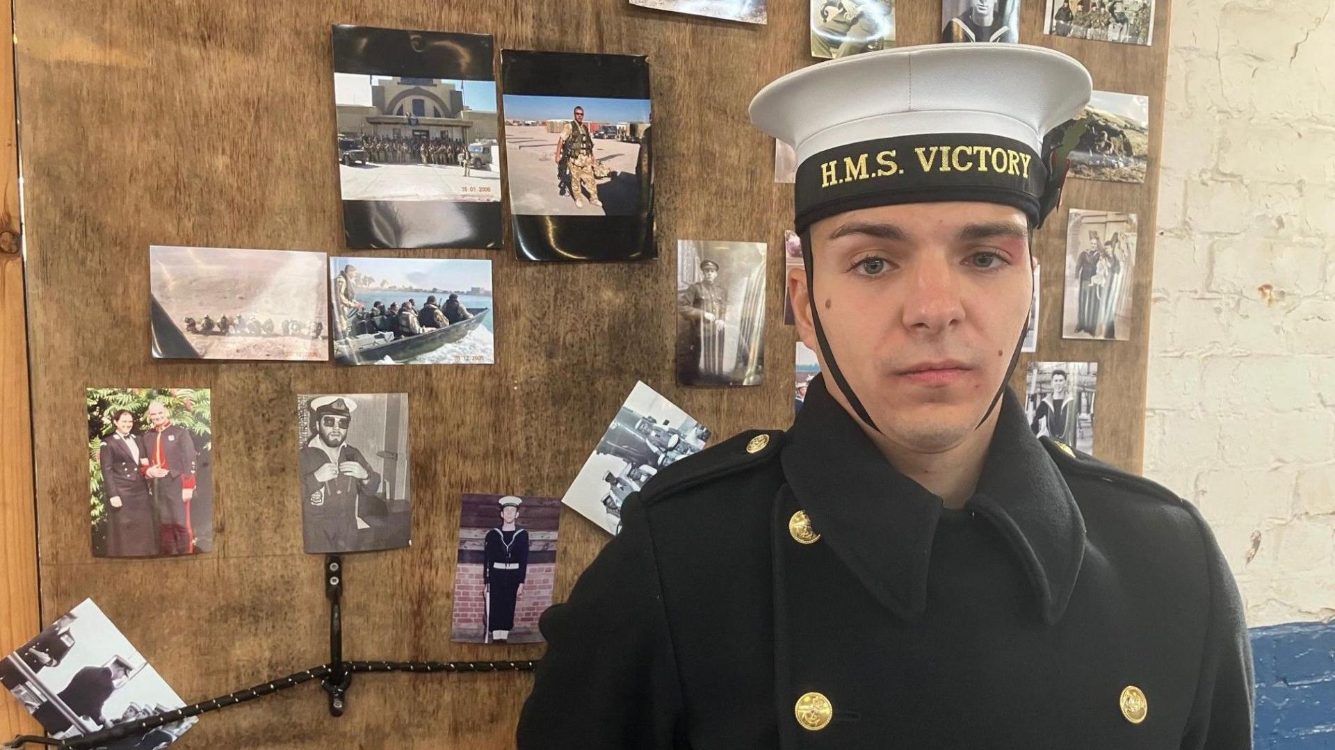 Able Seaman Jordan Reynolds stands in front of a board full of pictures of family members who have served in the military.