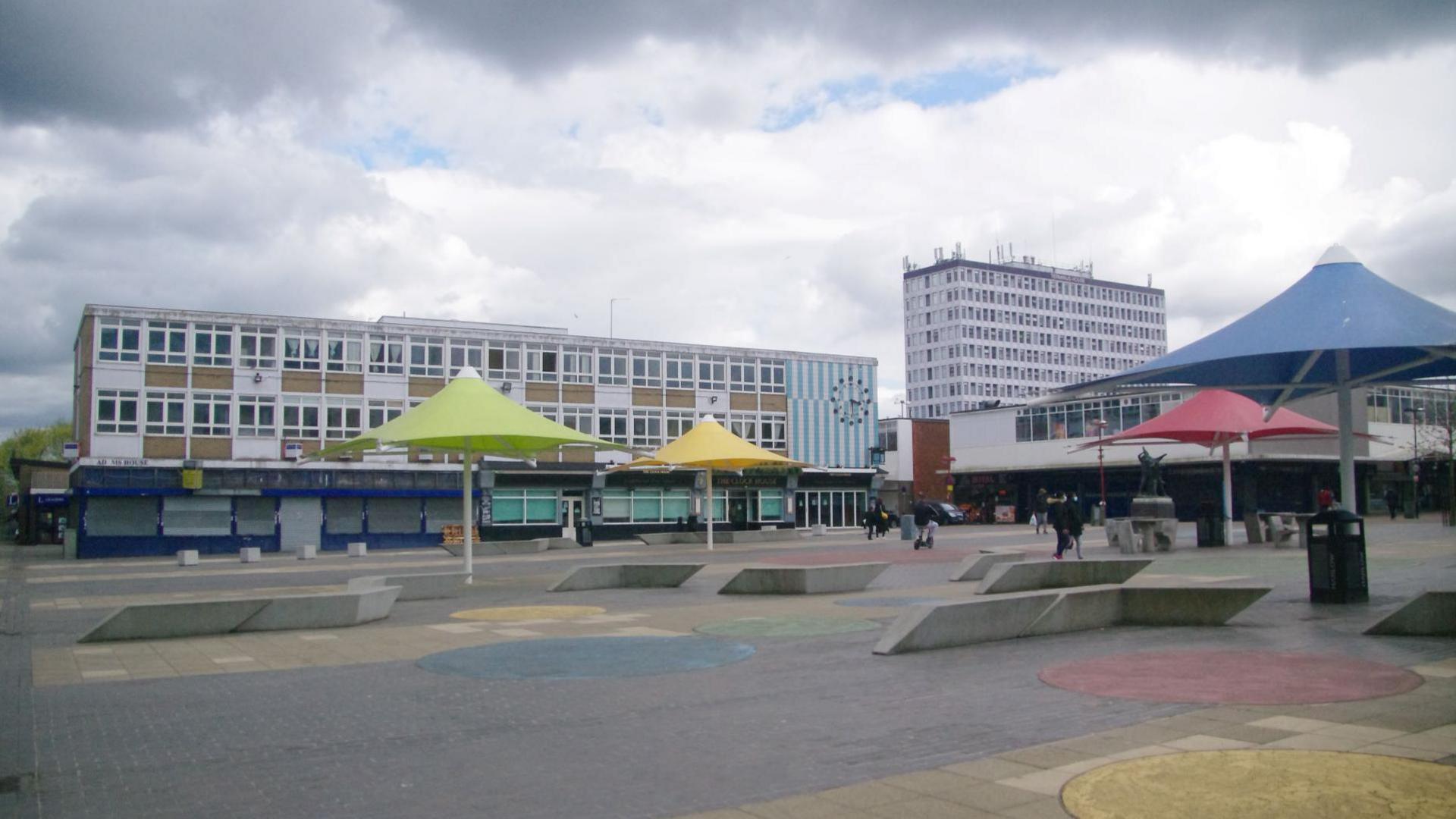 Market Square in Harlow, showing an open square with concrete seats, large parasols of different colours and various "Brutalist" style buildings in the background