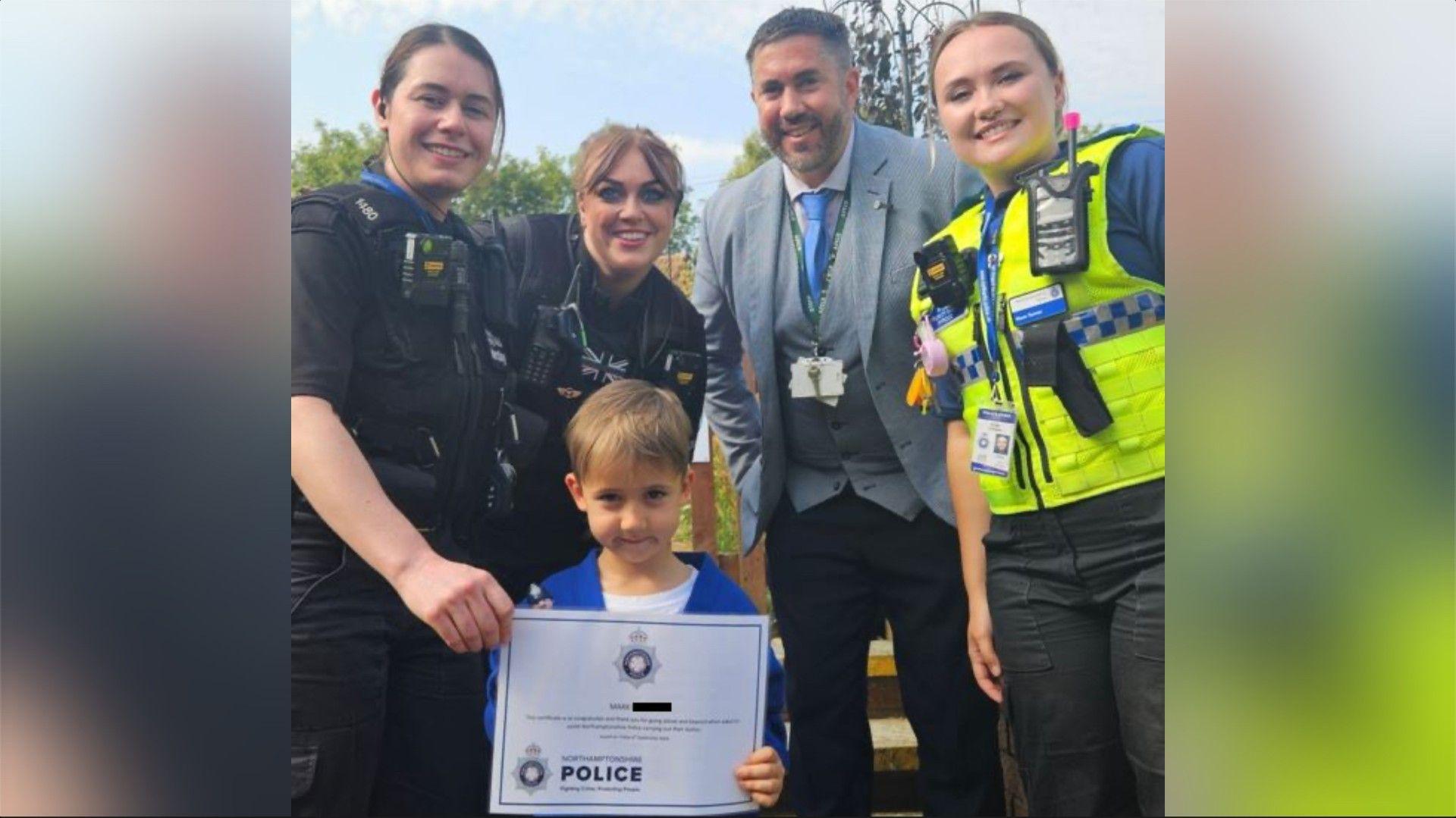 Three female and one male officer behind a boy wearing a blue top. Two women have black police uniforms and the other has a yellow hi-vis.  The man has a grey jacket and blue tie