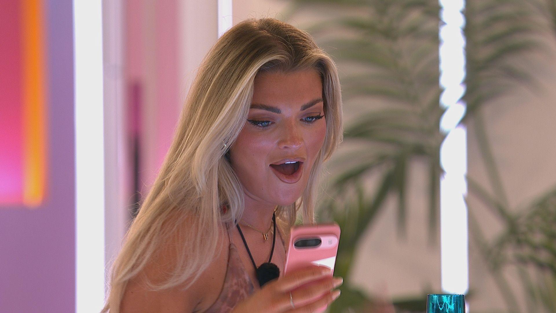 Islander Helena Ford looks at her phone with a shocked expression. She has long blonde hair worn loose and wears a pale pink strappy dress with a microphone around her neck. The villa in the background is decorated with palm leaves and pinks, purples and oranges.