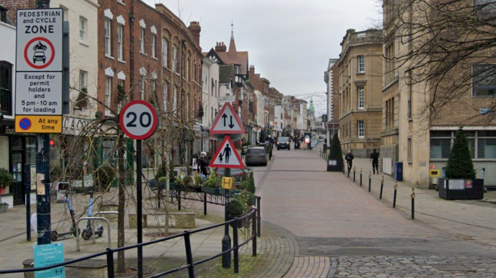 Gloucester city centre. The view facing up the high street. There are various signs for drivers including a speed sign and pedestrian warning. There are black bollards and a black railing to allow pedestrians to walk safely on either side of the street next to shops.