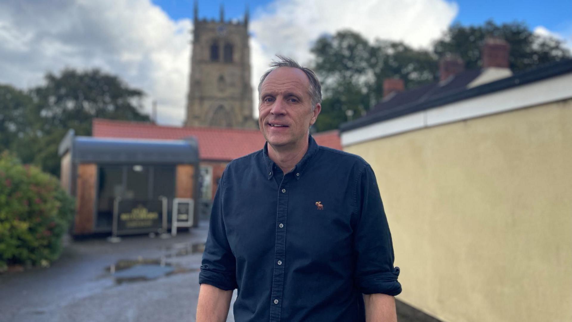 A middle aged man with closely cropped fair hair smiles outside a cathedral. He is dressed in a long sleeved navy shirt. There are puddles scattered along the pavement.