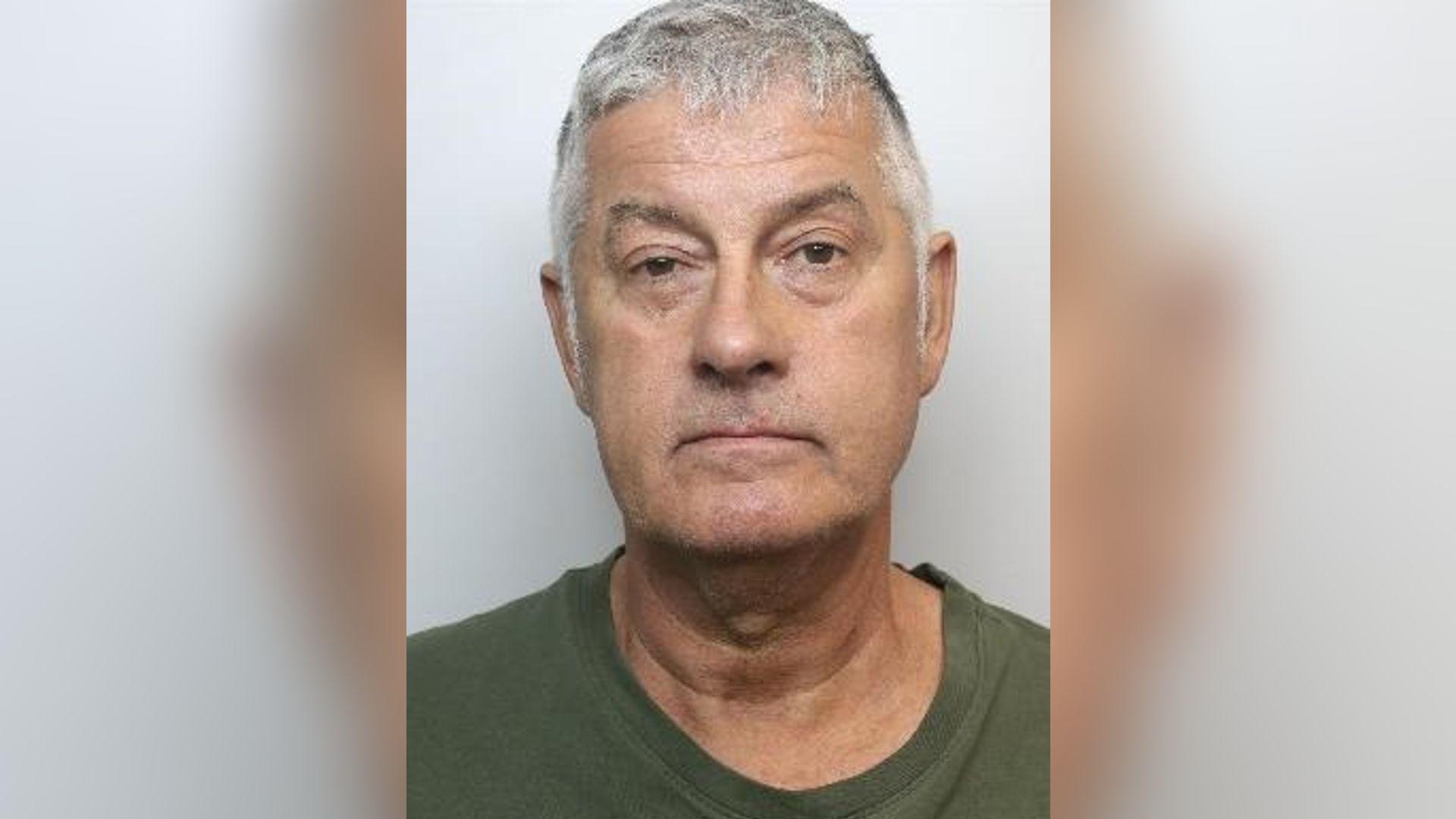 A police mugshot of a man with short grey hair wearing a green t-shirt.