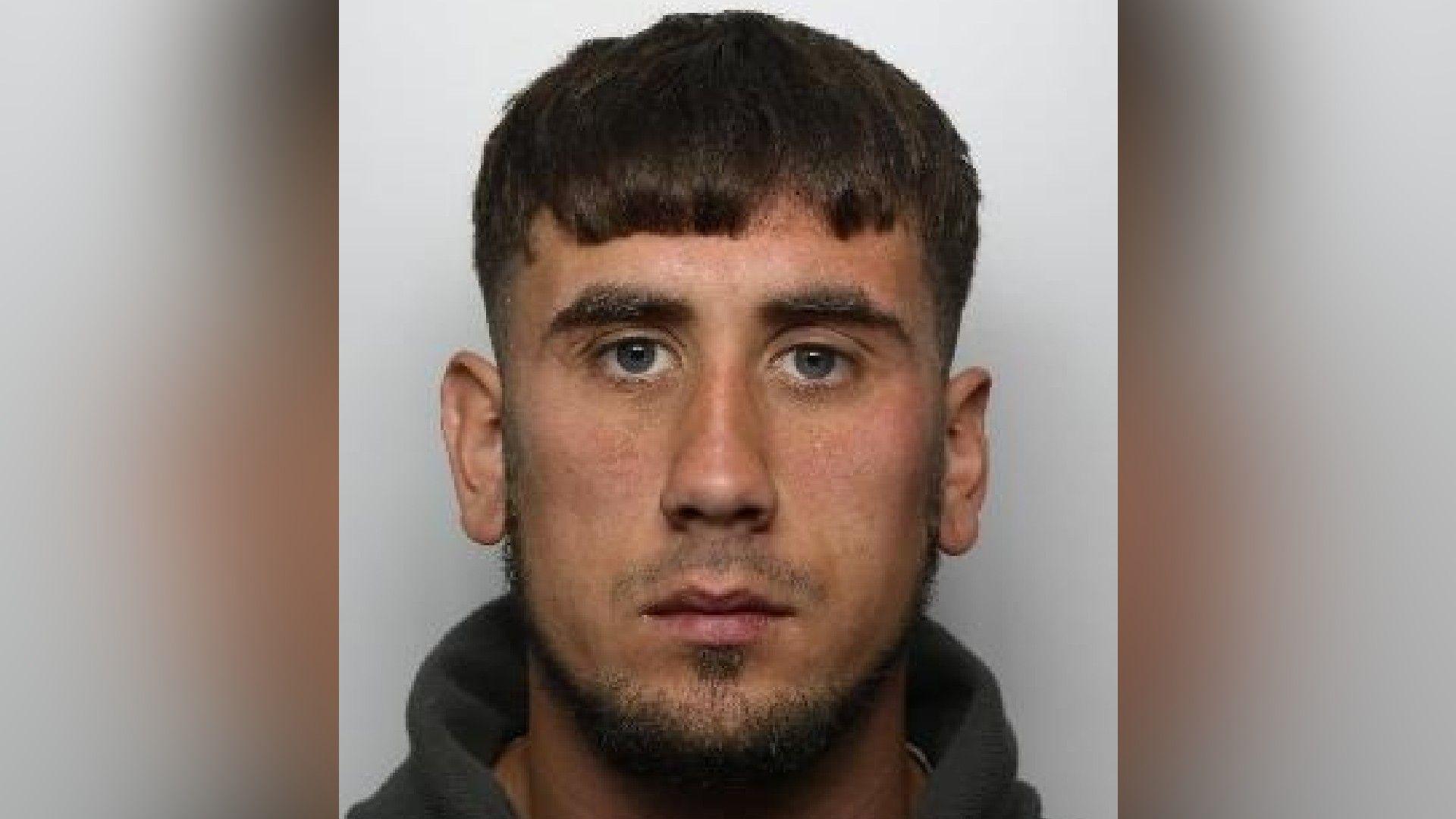 A mugshot of a man who looks straight at the camera. He has brown hair, combed over his forehead and facial hair. He is wearing a jumper.
