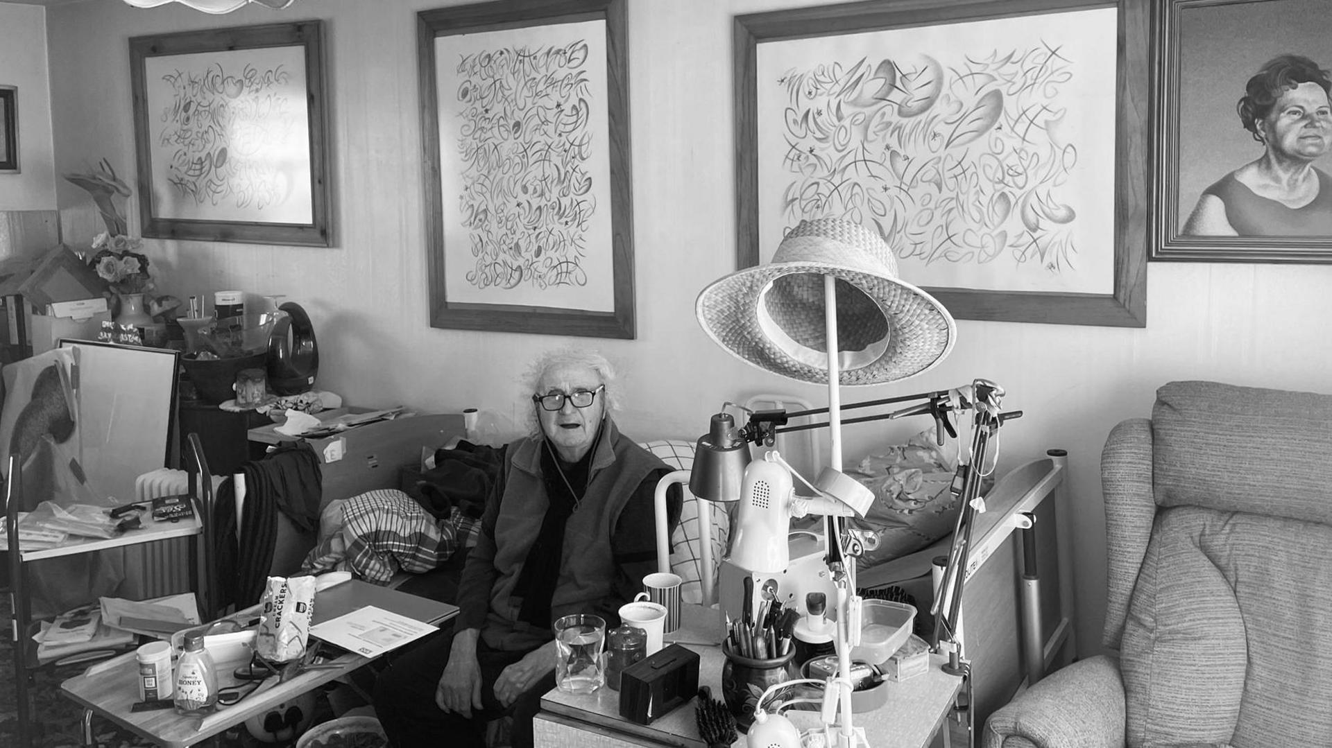 Black & white photo with Henry Orlik in his 70s in a living room with framed paintings behind him as she sits down, with a sofa to the right