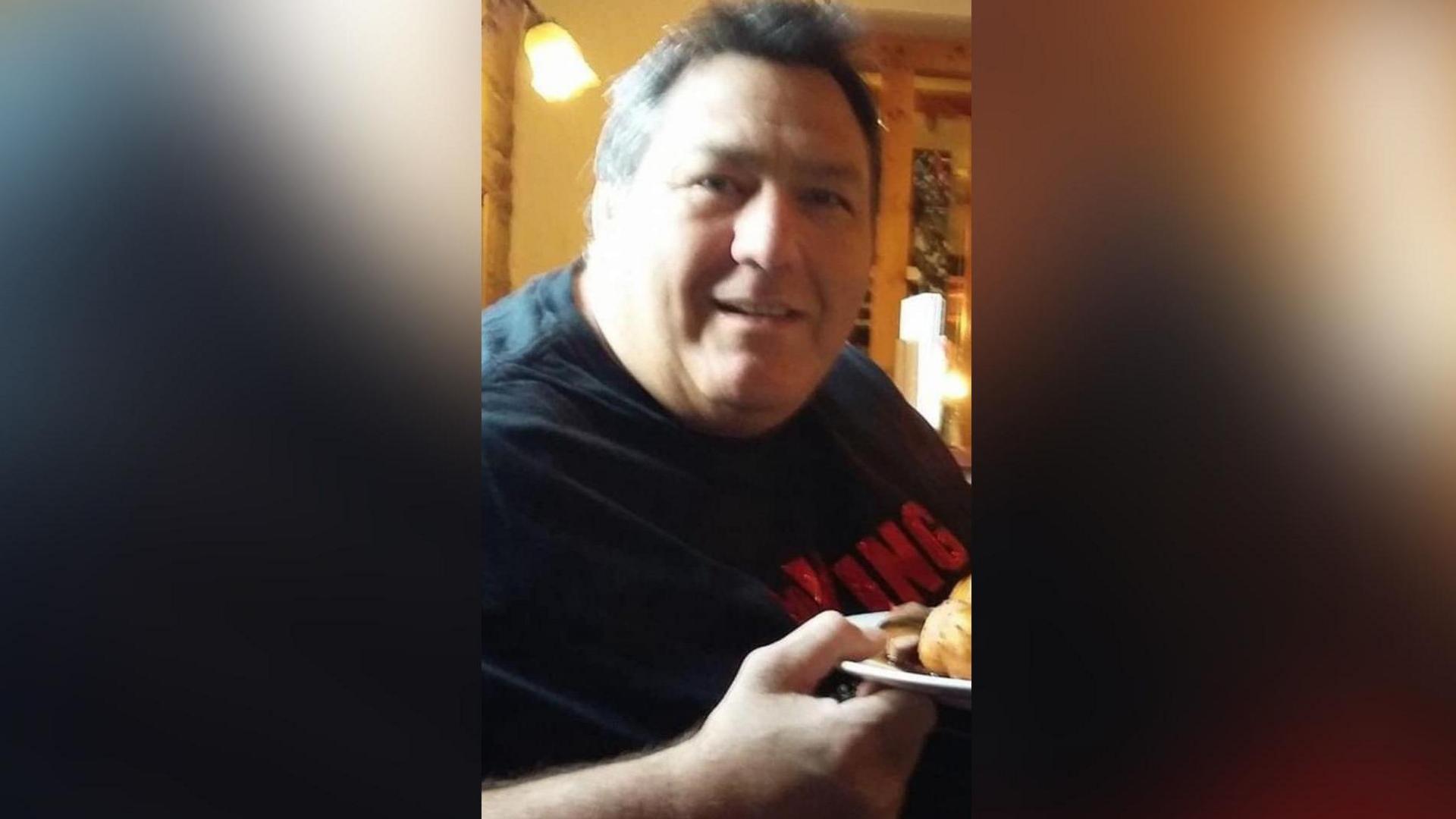 Mr Kirkwood holds a plate of food as he looks at the camera and smiles. He is wearing a black t-shirt and has grey hair.