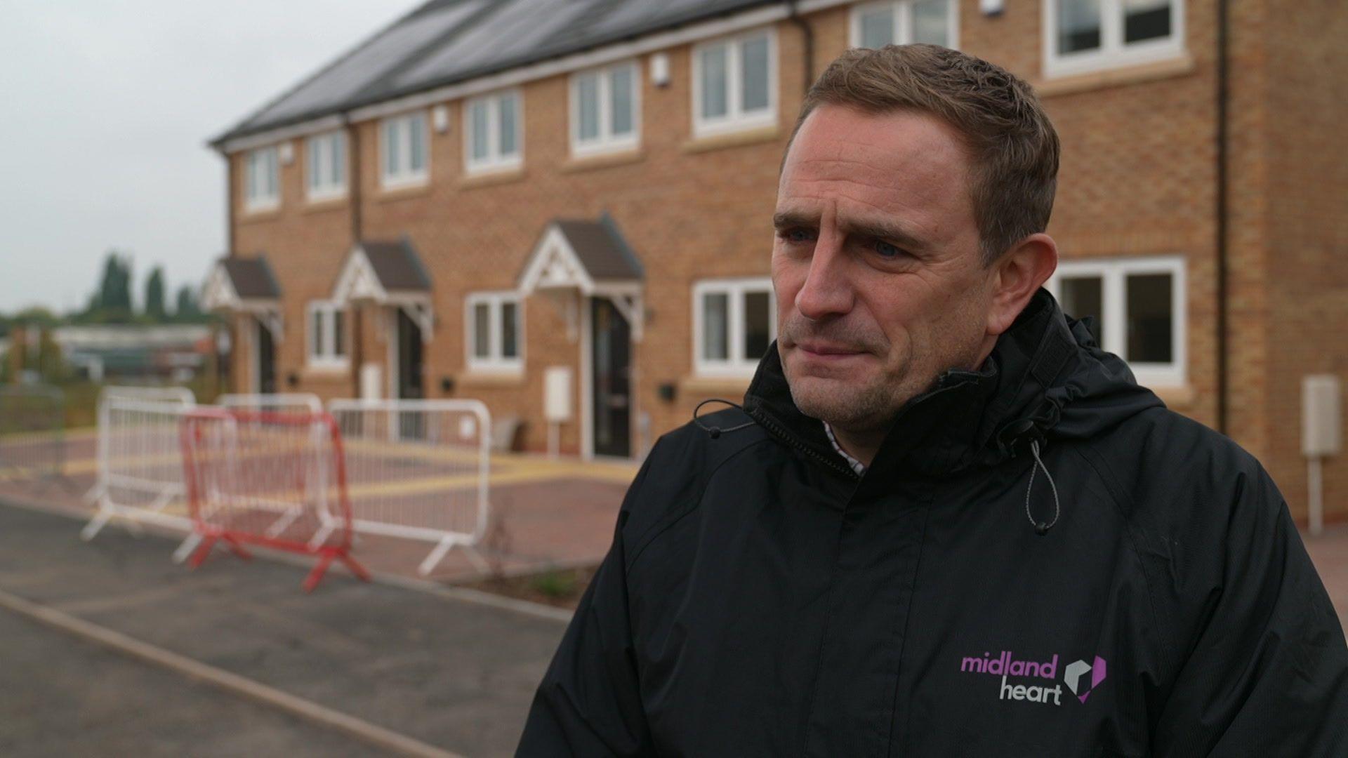 A man in a navy jacket with the logo Midland Heart in purple and white stands in front of a row of red brick houses that have just been built 