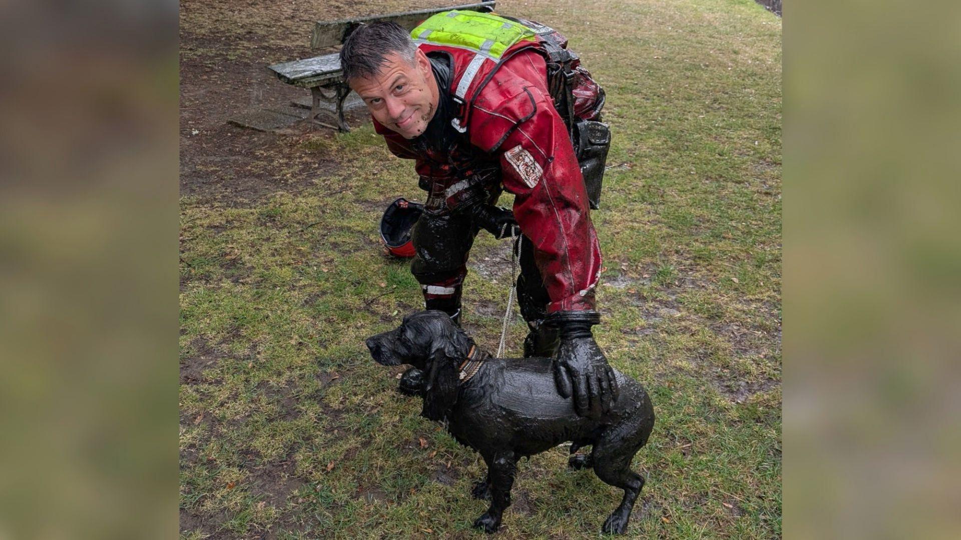 A muddy Bailey is standing looking away from the camera with a firefighter holding the lead. They are standing on a patch of grass near some woodland. Bailey is covered in mud and still looks a bit startled. The firefighter is crouched slightly with his hand on Bailey's back and looking at the camera. The man has short greying hair and wears a waterproof red all in one uniform, with a high vis patch on the back.