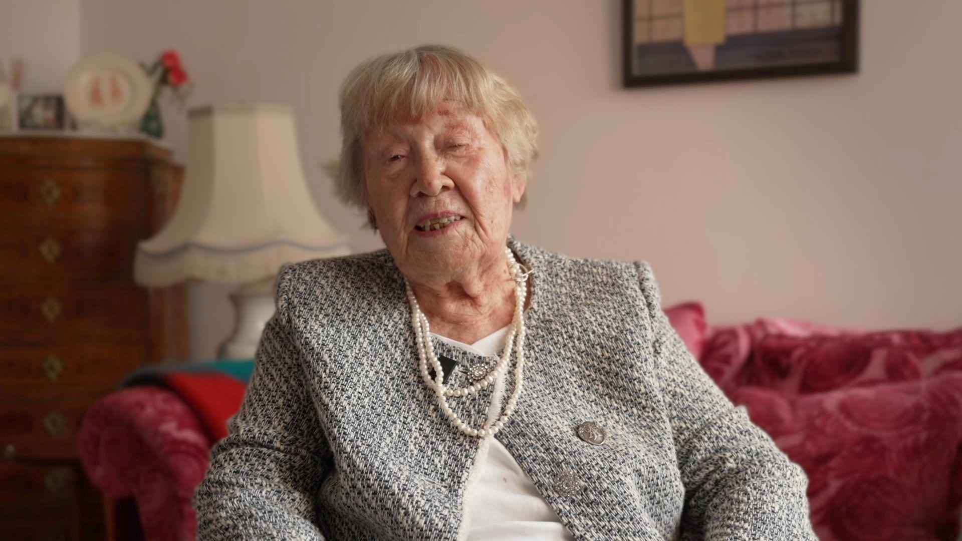 Audrey has short blonde hair, she is wearing a grey blazer with a pearl necklace and is sitting in front of a red sofa with a white, fringed lampshade behind her.