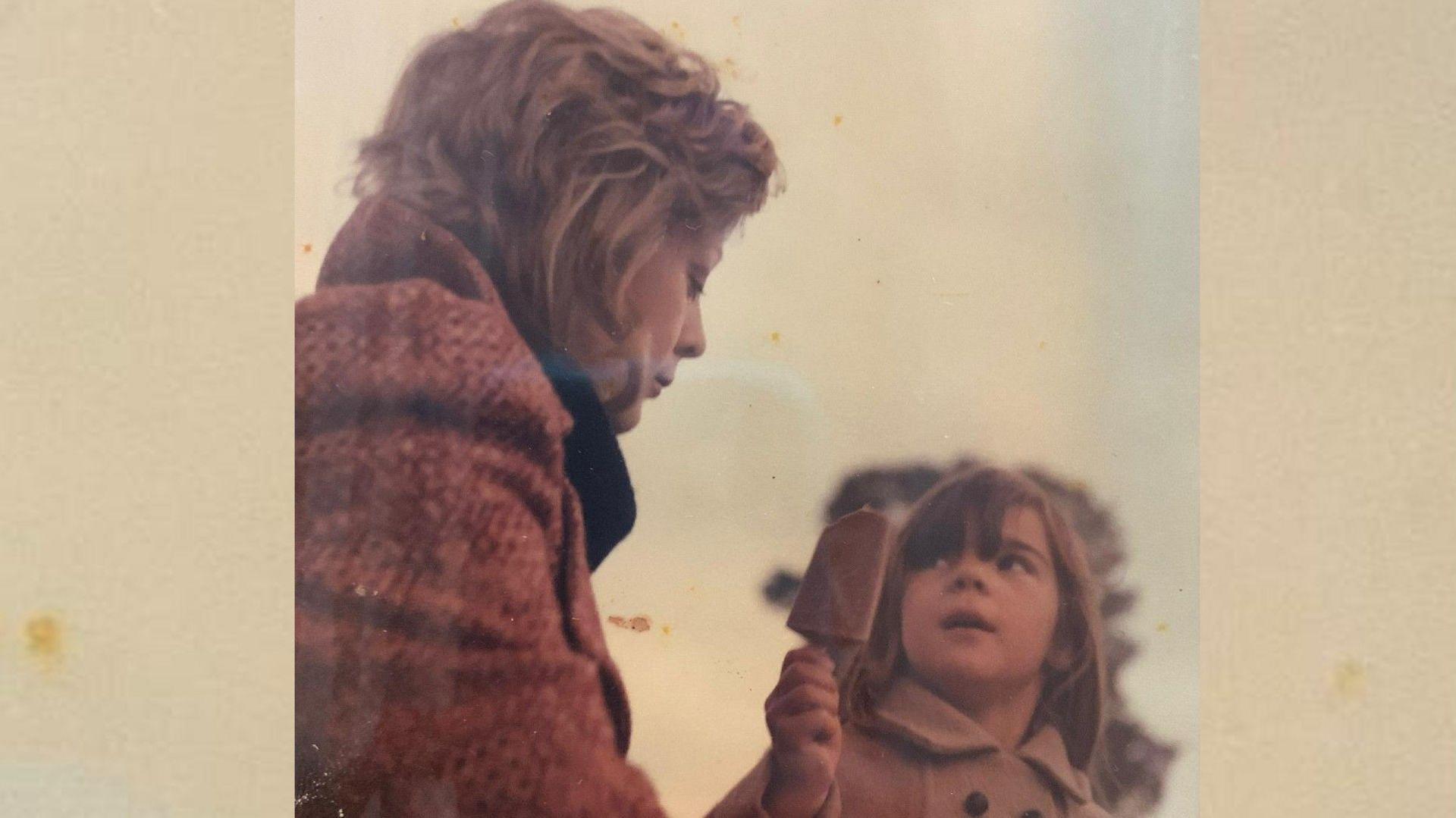 An archive photograph of a little girl wearing a coat holding an ice cream. She is looking at her mum who is also wearing a coat. They are sat in a park.