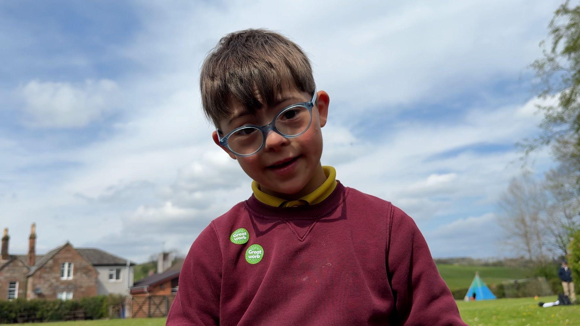 Young boy wearing a maroon jumper stands in a garden.