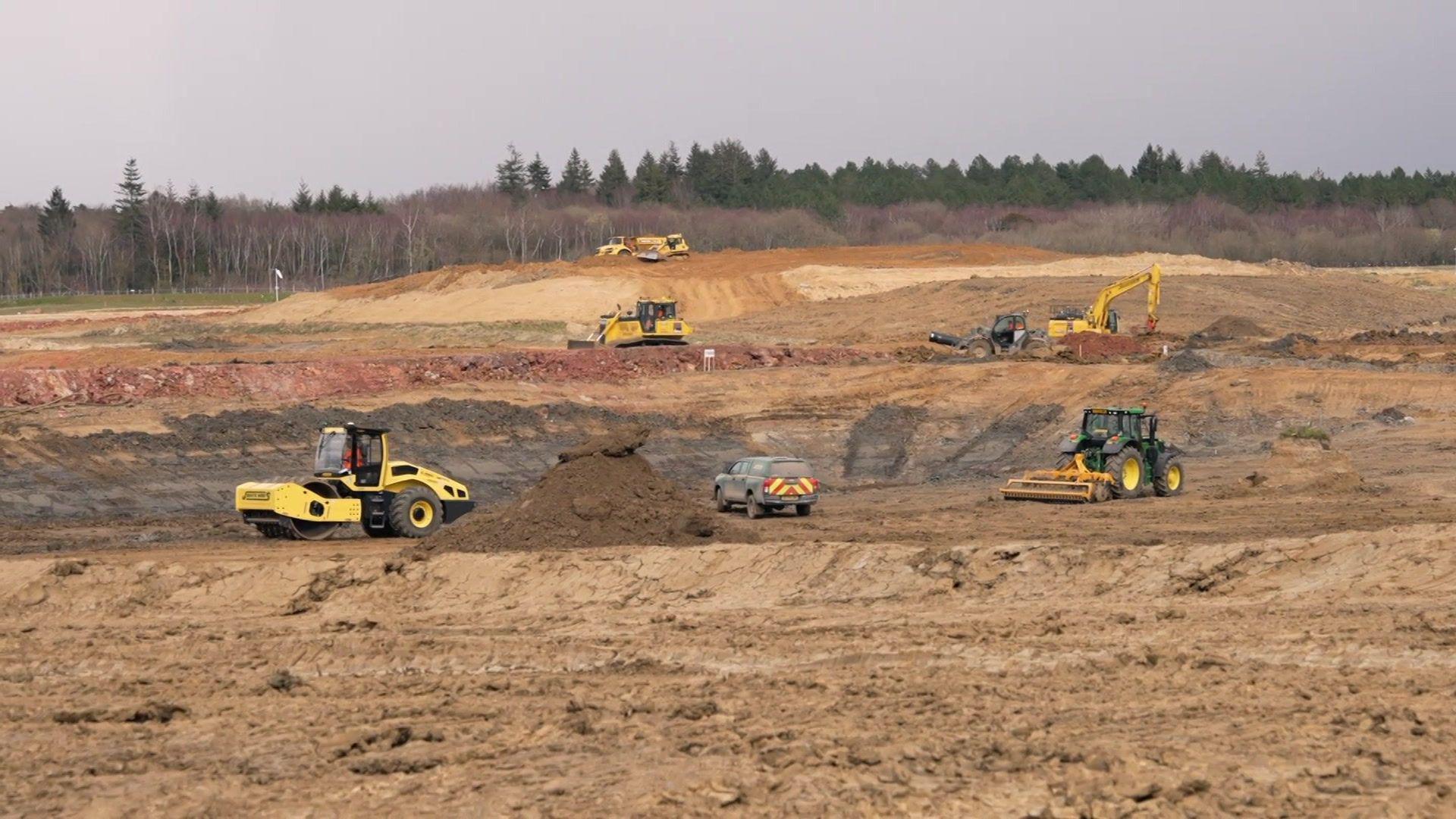 A section of land where the Havant Thicket Reservoir is being built. The land is brown dirt and there is some hillside in the background and a forest of trees. There are yellow diggers dotted across the landscape and a 4x4 truck.