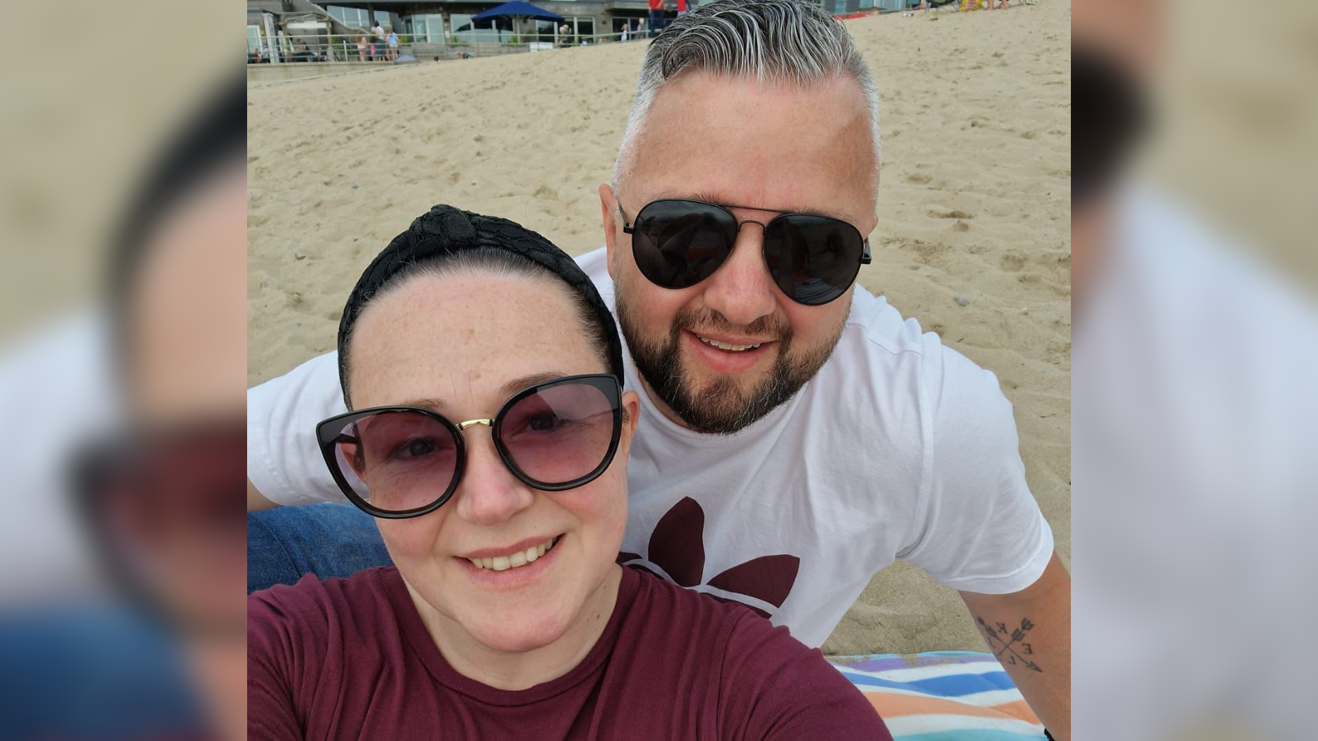 Ed and his wife Kerry sitting on a beach smiling. Kerry is sat on the left and is wearing a maroon tshire, sunglasses, and has dark tied-back hair. Ed is on the right, has grey hair, is wearing sunglasses and a white tshirt.