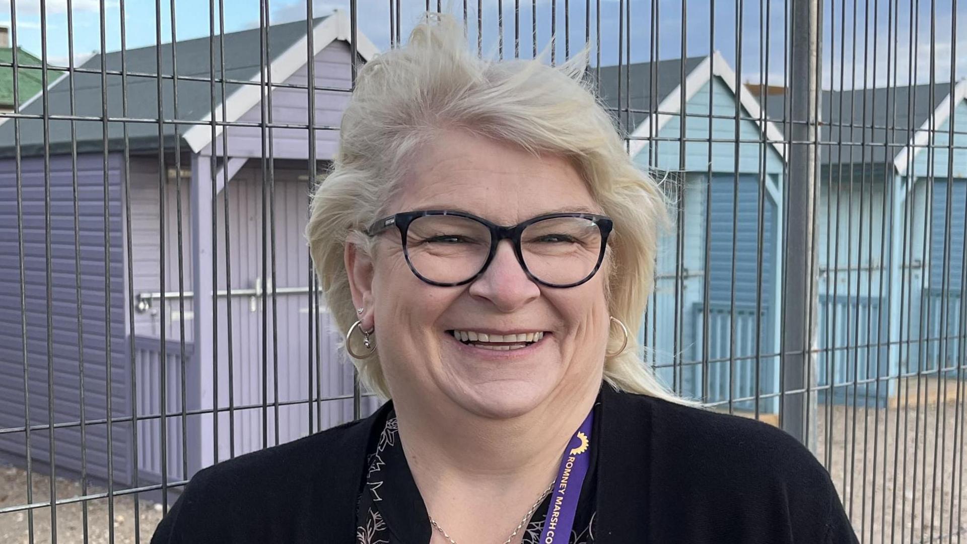 A blonde woman with short hair and black glasses smiling at the camera. She is standing in front of a row of purple and light blue beach huts.