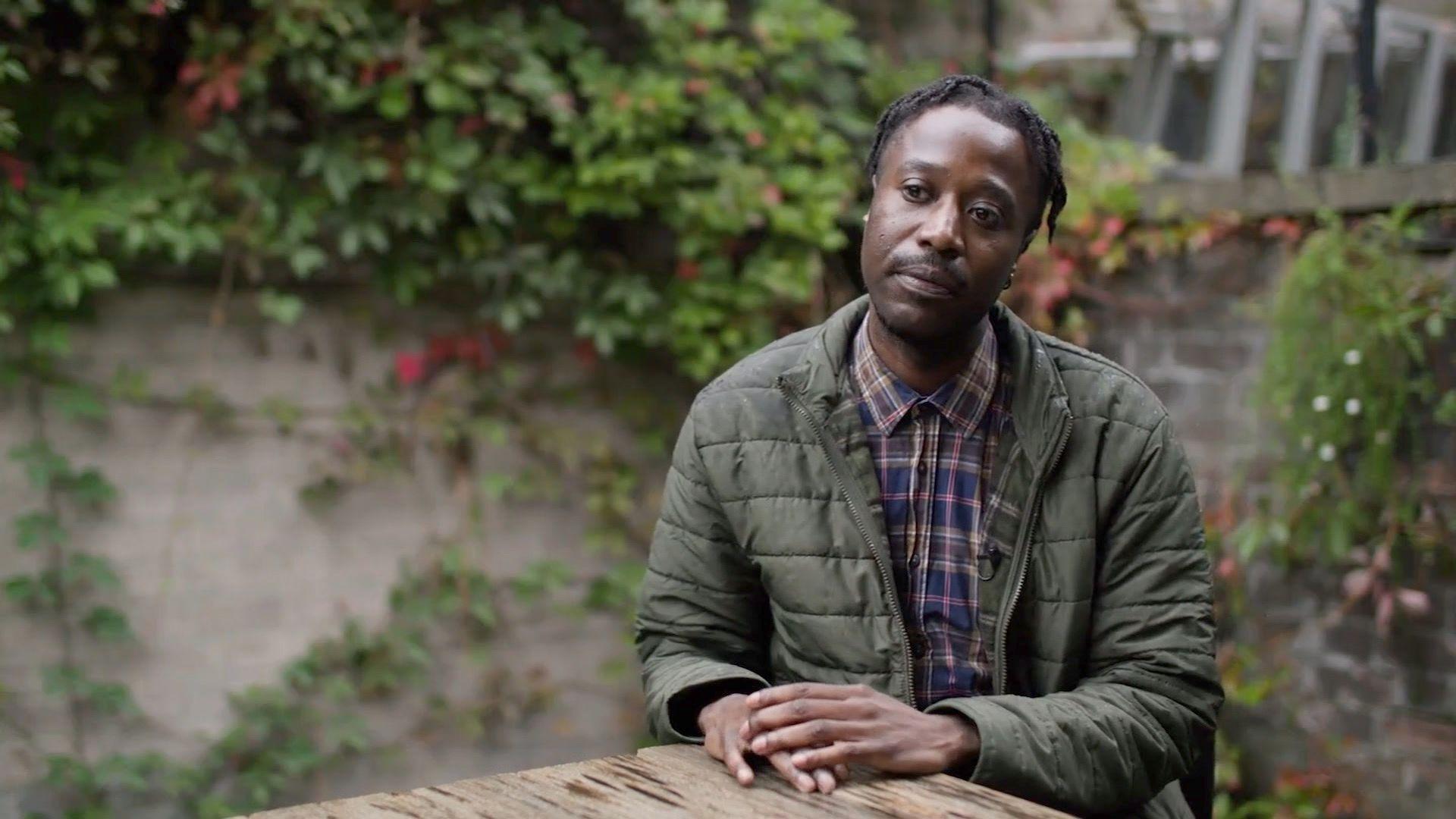 Jeanperry Mundabi is sat outside at a wooden table, in front of a wall covered in plants. He is wearing a green coat and purple checked shirt.