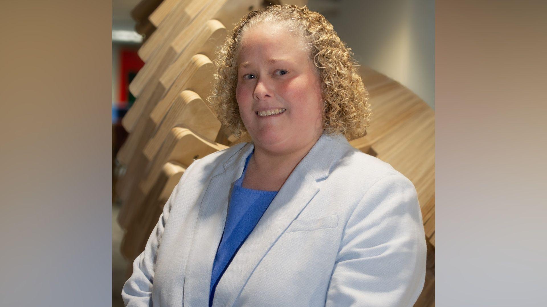 A professor with a very curly blonde bob smiles. She is wearing a white doctors coat with a blue top underneath and there is a wooden sculpture behind her.
