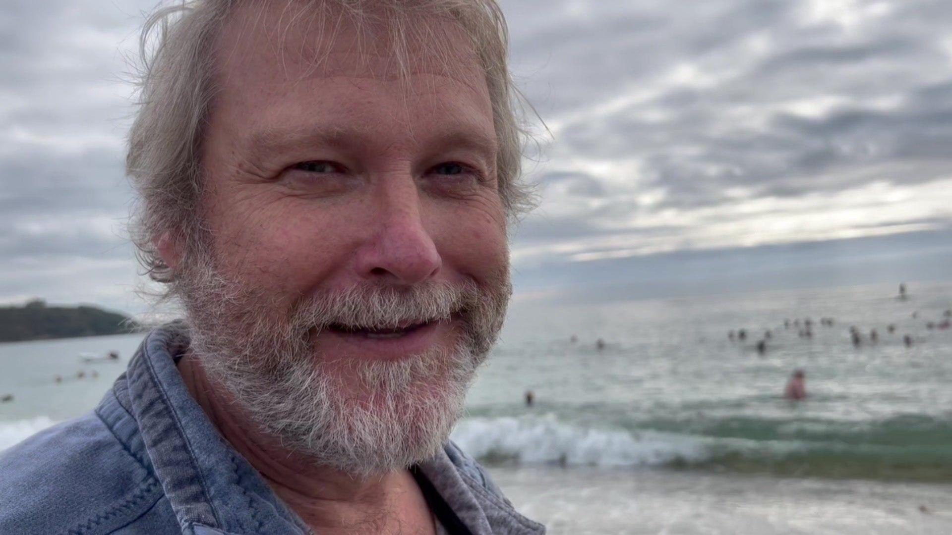 Dave Hitchcock, Ruth's husband, standing on the beach watching all the swimmers who have gathered to celebrate her achievement. He has grey hair and a beard, and is smiling at the camera.