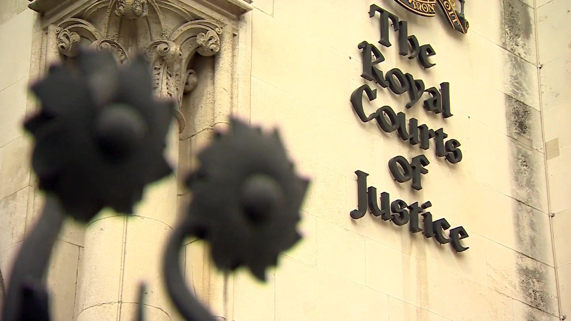 The stone facade of a building with "The Royal Courts of Justice" displayed in black lettering on the wall. There are floral iron decorations from the top of a gate in the foreground.