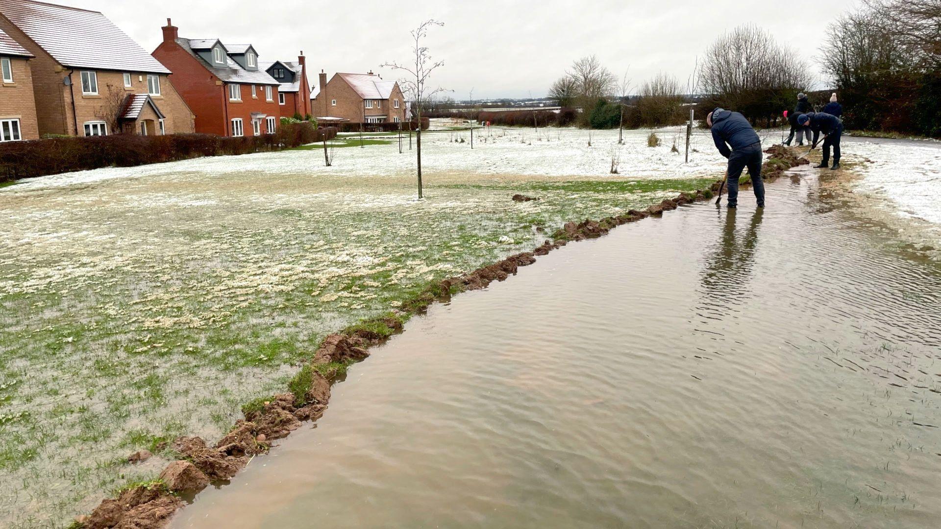 A trench filled with water can be seen with three residents digging. A waterlogged lawn can be seen to the left of the trench with a row of houses to the far left.