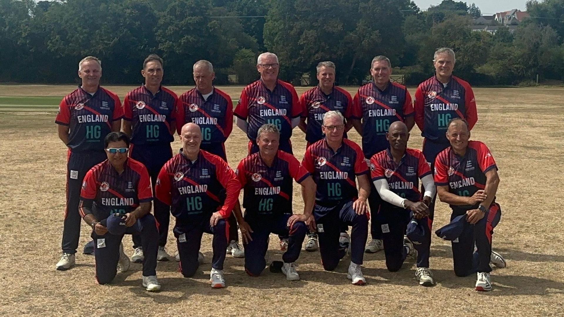 The 2026 England Over-60s World Cup squad looking towards the camera with barren grass in the foreground and green trees in the background.