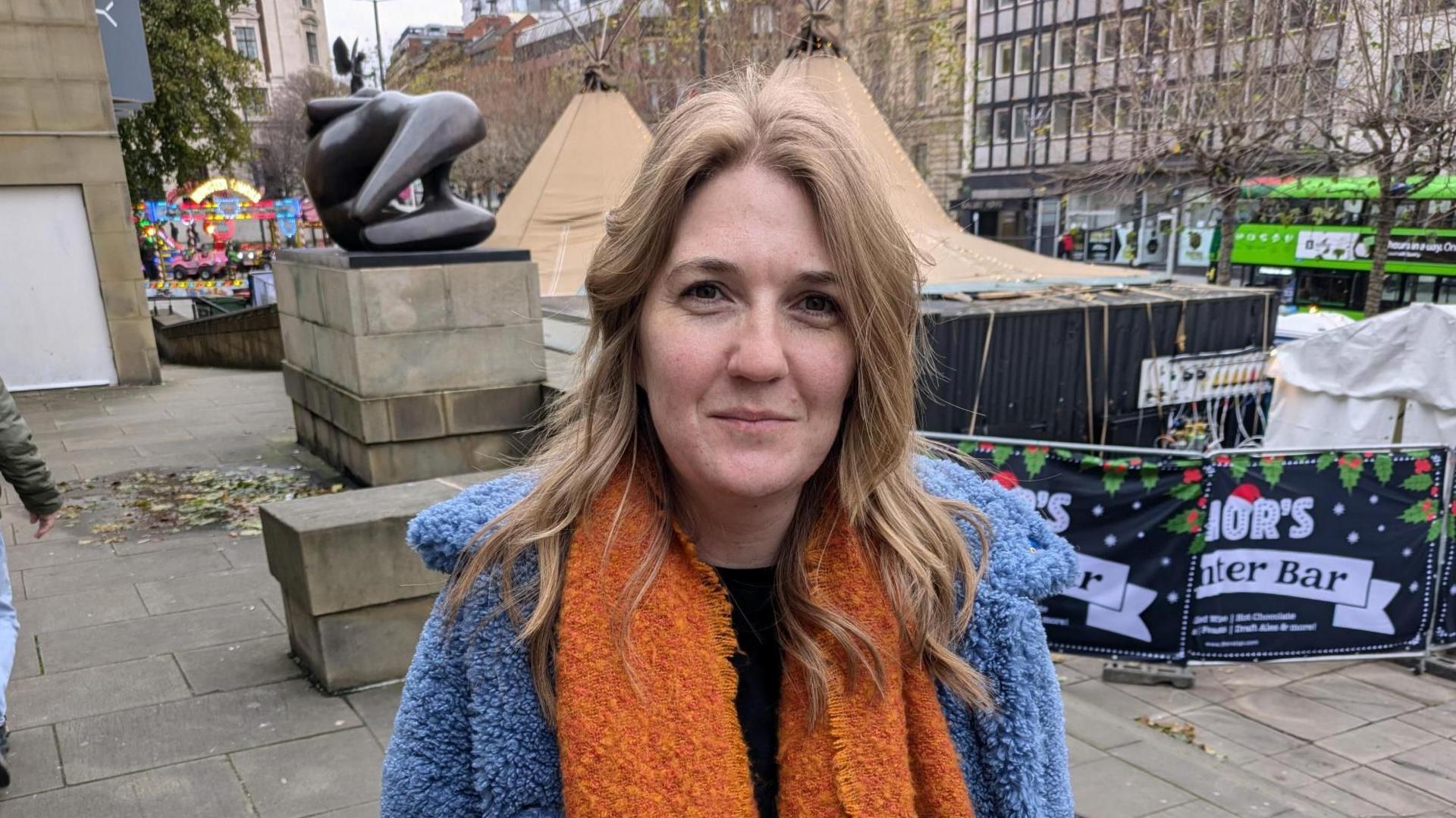 A woman in her thirties with fair hair, a blue coat and an orange scarf. She is smiling into the camera.