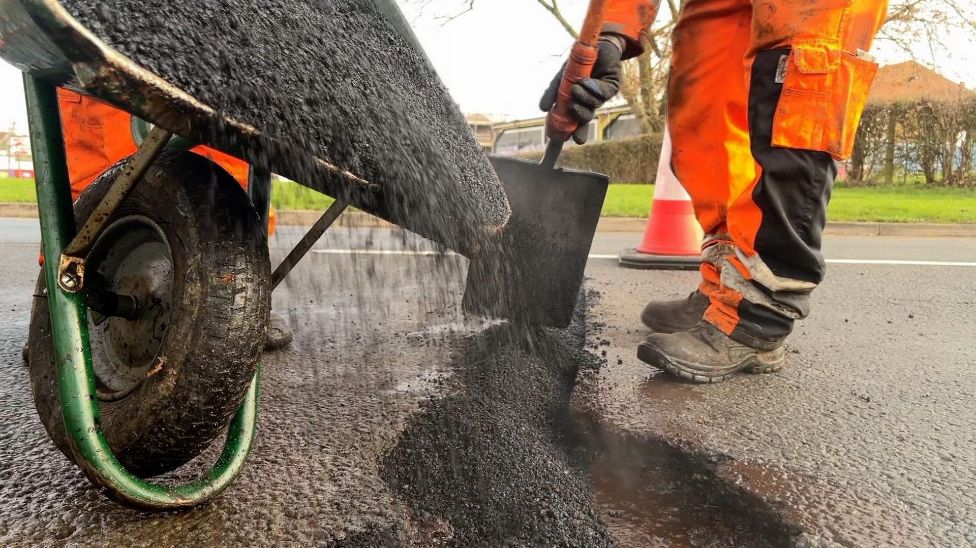 A wheelbarrow tipping gravel into a large pothole on a road in Swindon while a roadworker in an orange boiler suit spreads the material with a spade