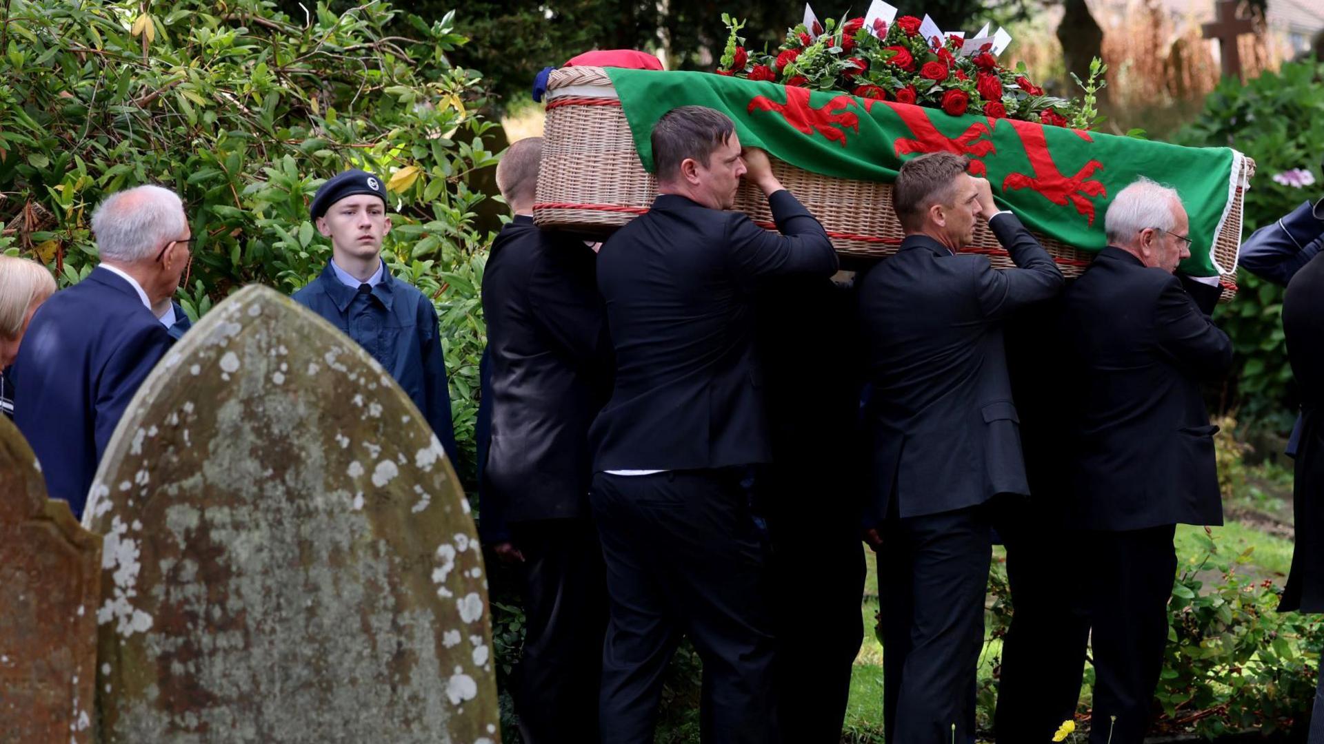 Hefin David's coffin draped in a Welsh flag with red roses on the top, being carried by six people, with three men seen from the side. A young air cadet can be seen to the coffin's left.