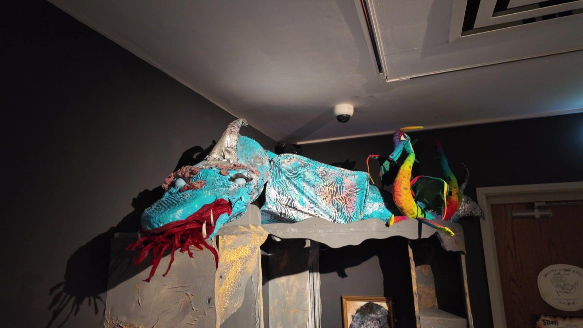 A big blue dragon with red paper flames coming out of its mouth placed on top of a shelf at the Story Museum. A smaller rainbow-coloured dragon artefact is standing on its back feet next to it. The room is dimly lit to represent a cave.