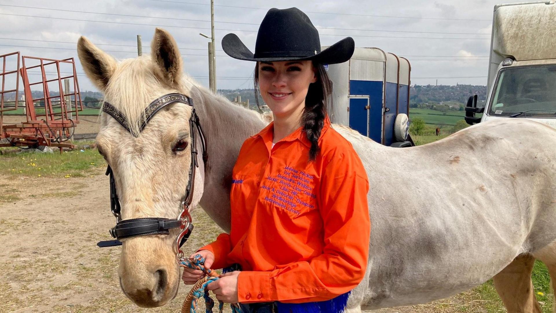 A teenage girl wearing a bright orange shirt and cowboy hat stands smiling next to a grey pony.