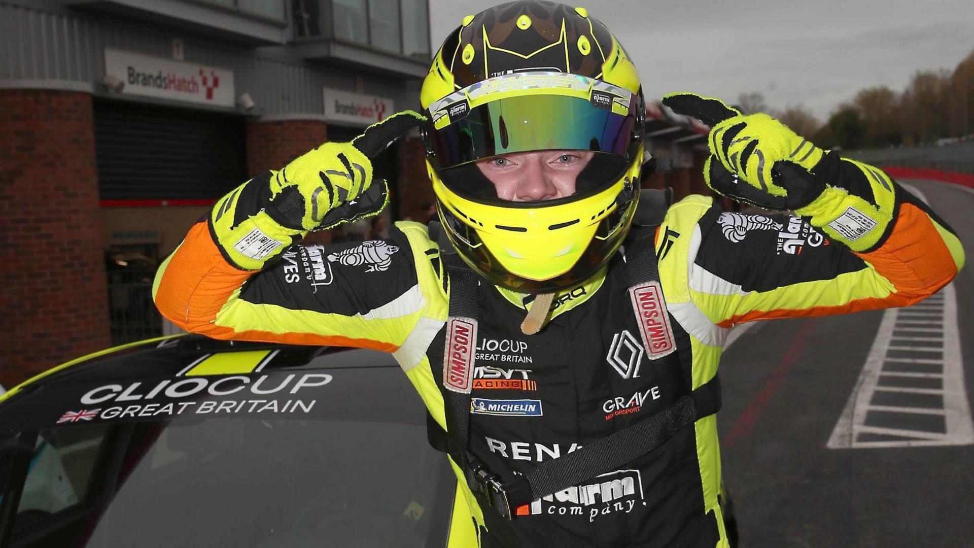 Nicky Taylor pointing with his index fingers to his head. He is wearing yellow, black and orange racing overalls and is standing in front of his Clio on a racetrack.