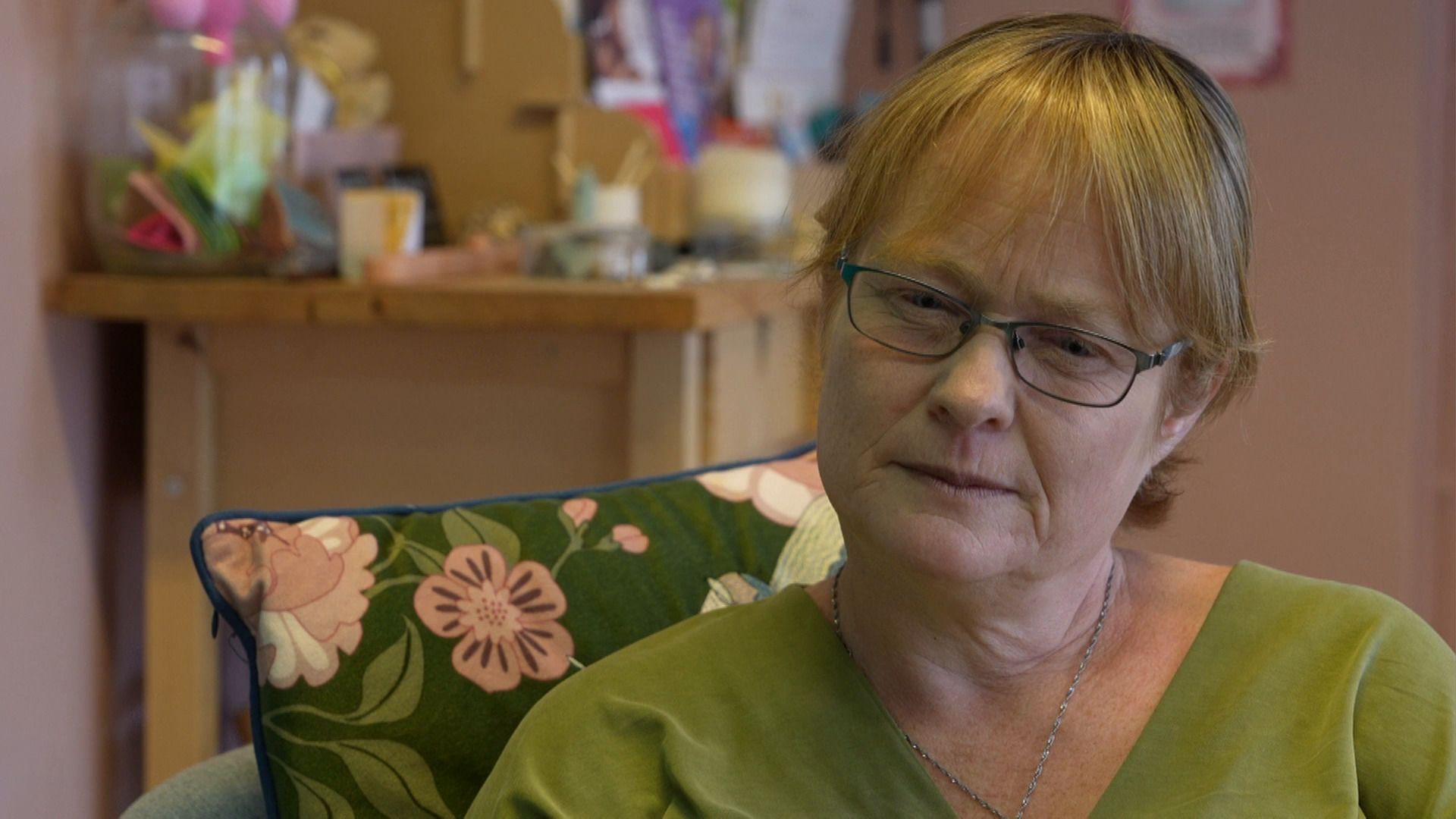 Katherine Elliot who is wearing blue glasses, she has short ginger hair, with a green shirt, sitting behind a green and pink floral cushion.
