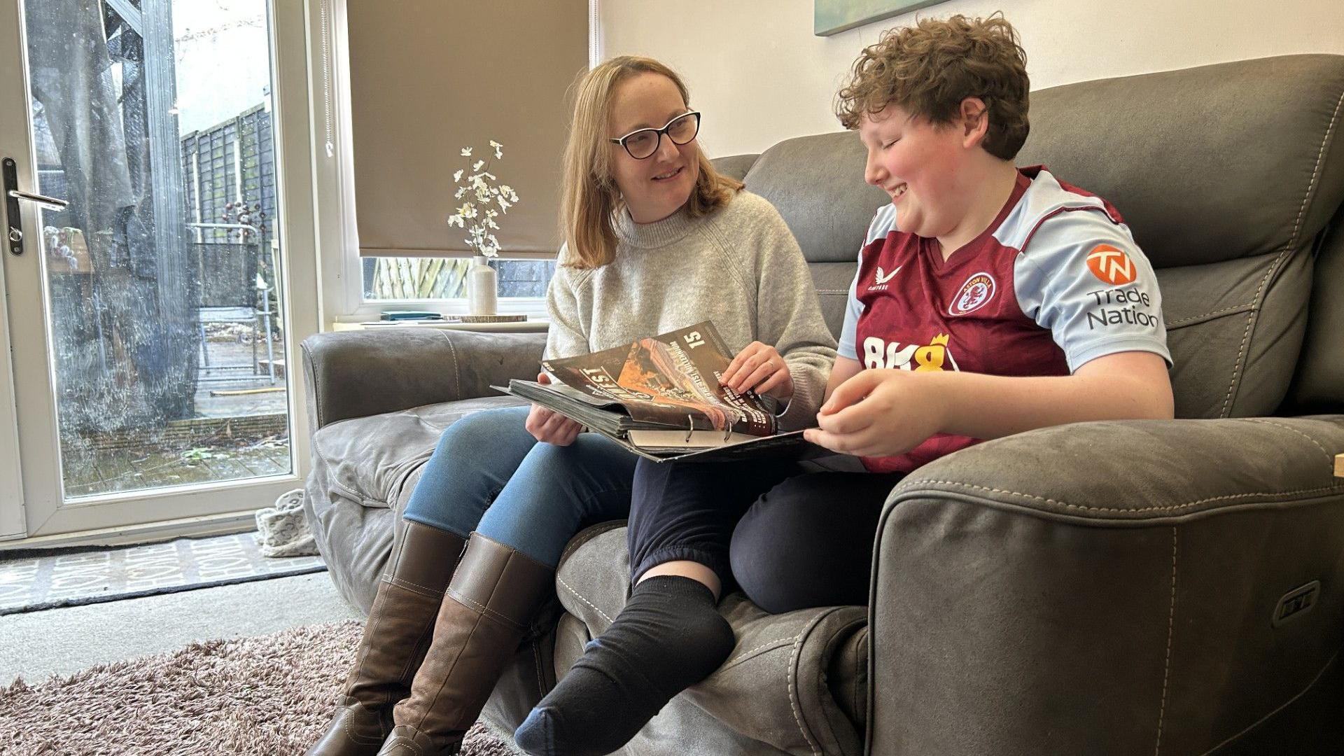 Amy wears glasses, a grey jumper and long brown boots. She is sat next to her 12-year-old son Cooper, a child who has brown hair and is wearing an Aston Villa top. They are sitting on a sofa in a living room looking at a book with a French door behind them. They are both smiling.