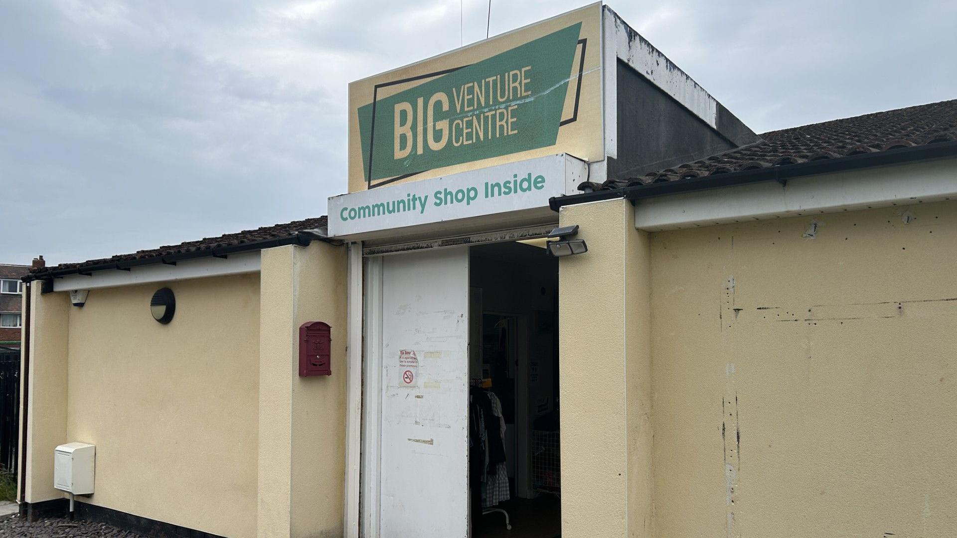 The Big Venture Centre is a single-storey yellow building. Some of the paint has chipped away. One of its white doors also looks like a little old and tired. The sign says: Big Venture Centre. Community Shop Inside.