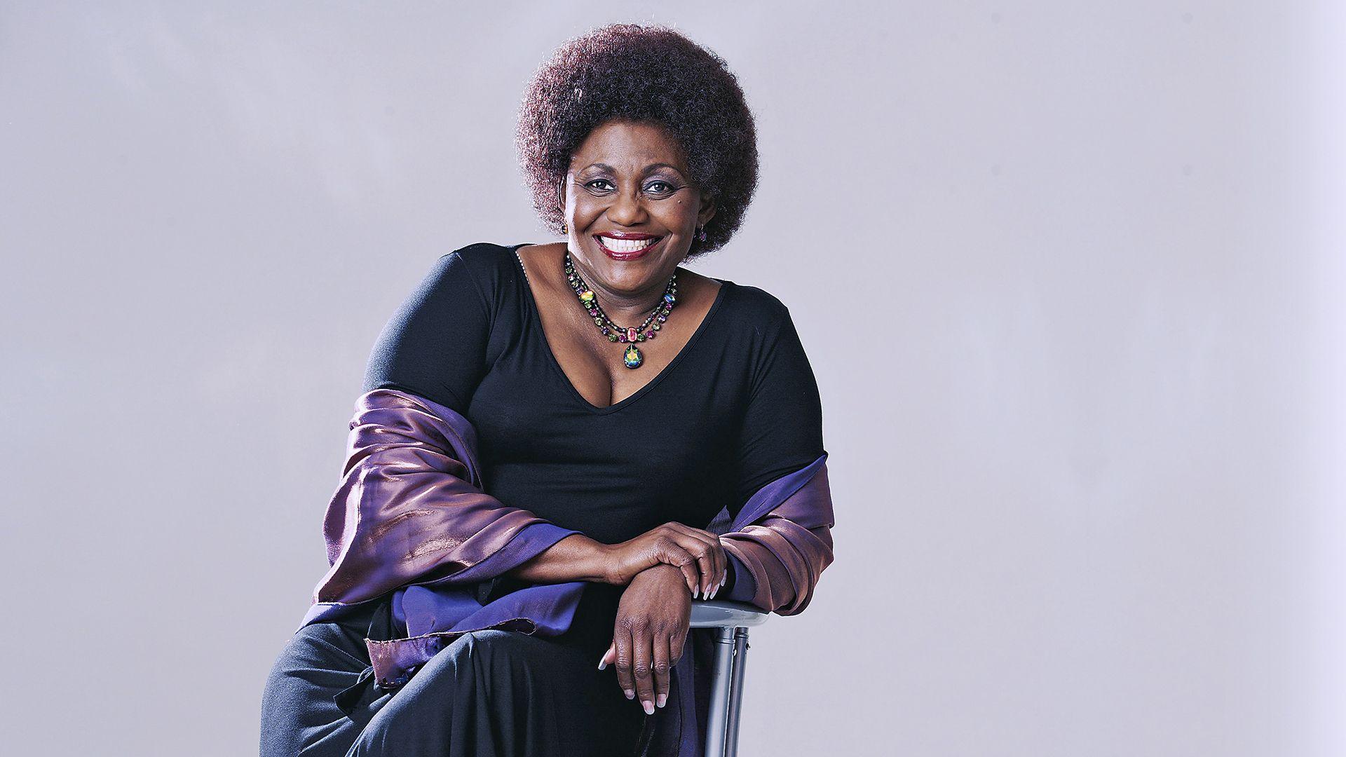 A portrait style image shows actress Nandi Nyembe posing for the camera while smiling and sitting on a chair in a photography studio against a grey background, taken in South Africa in 2007.