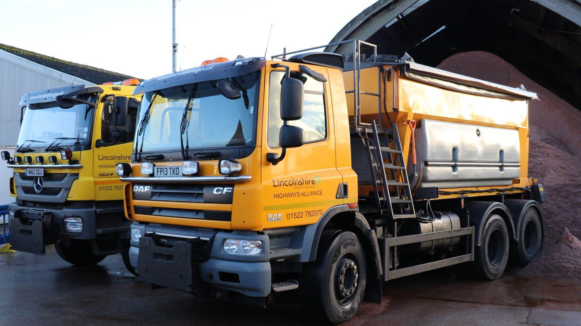 Two Lincolnshire County Council highways gritters parked in front of a large pile of gritting sand. Both vehicles are yellow and have council highways signage on the passenger side door