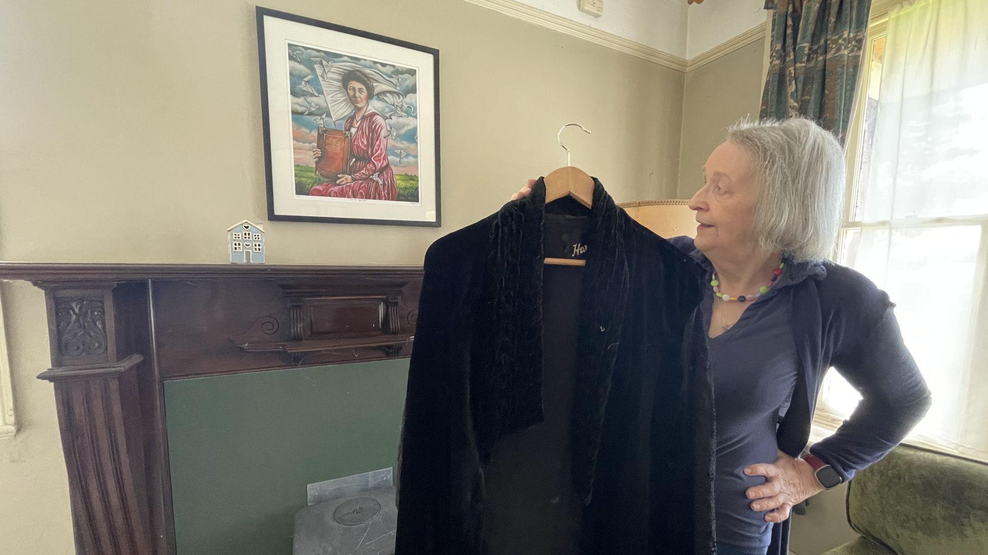 Meg Ellis yn dal y clogyn ac yn edrych ar bortread yr artist Meinir Mathias o'i mam-gu